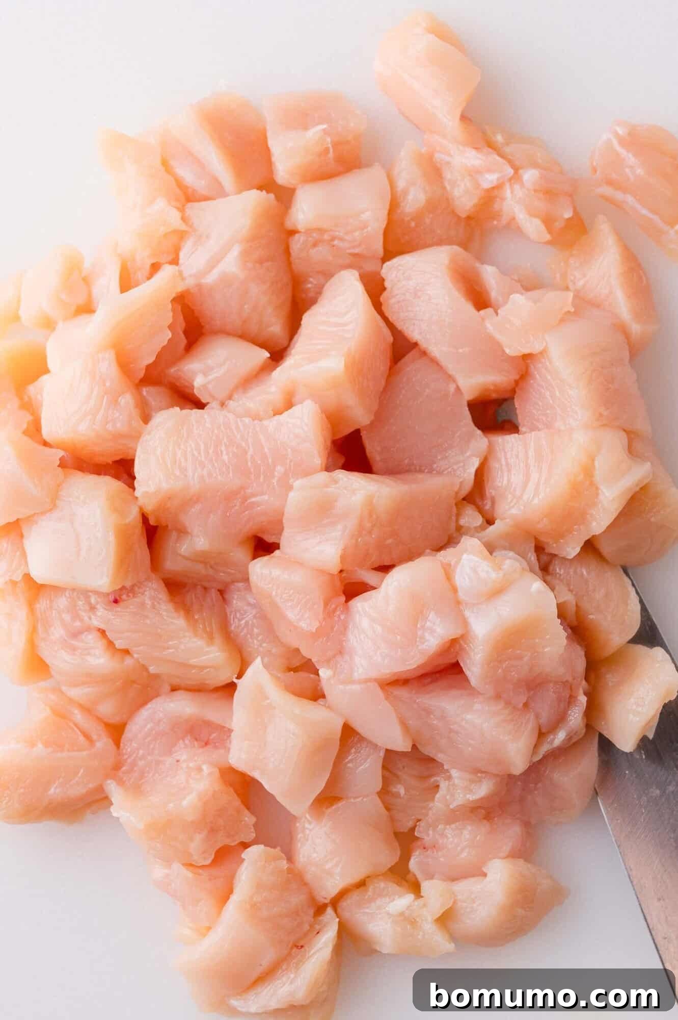 Diced raw chicken pieces on a white cutting board with a knife visible beside them.