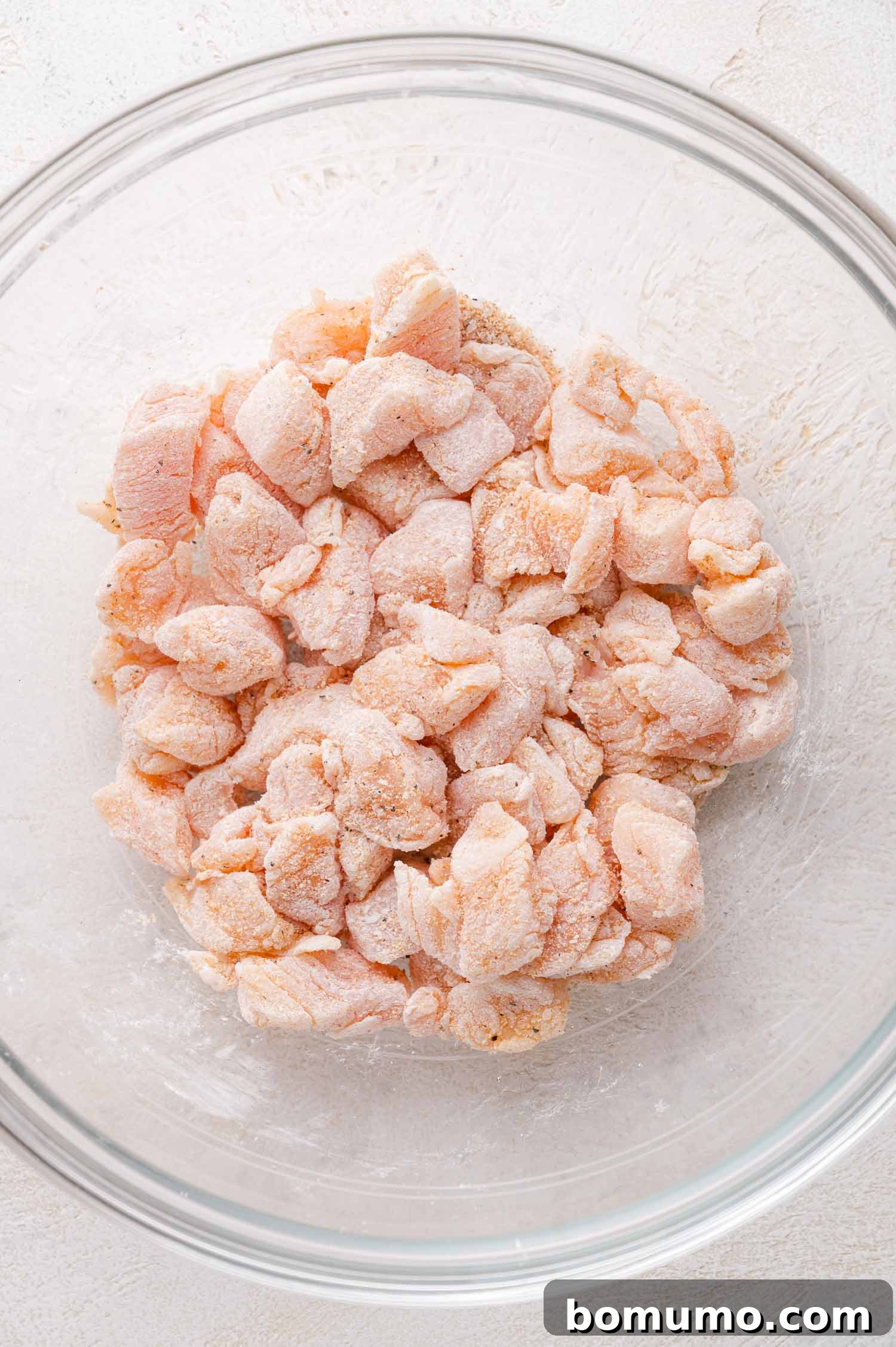 Raw, cubed chicken pieces coated in a light layer of cornstarch and seasonings in a clear glass mixing bowl on a white surface.