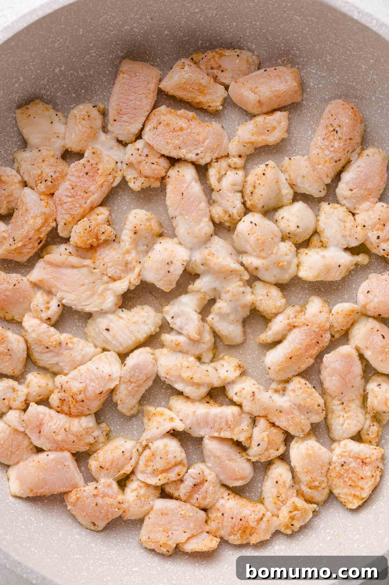 Cubed pieces of seasoned chicken breast cooking in a nonstick pan, lightly browned on the edges.
