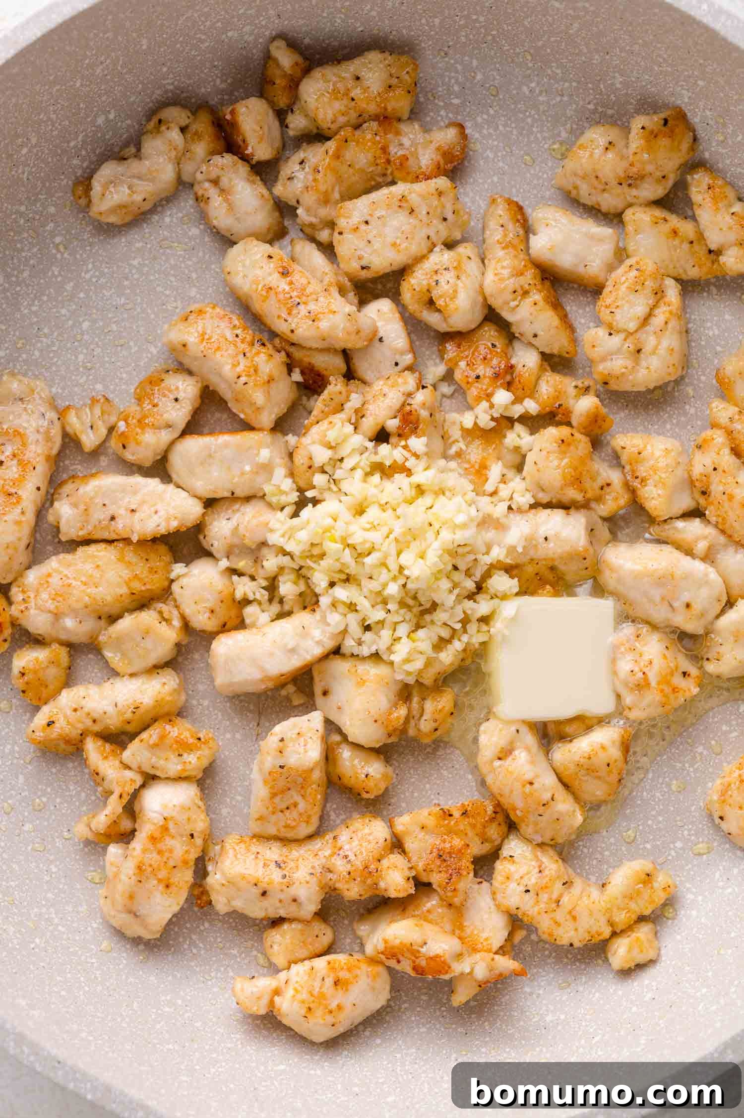 Cubed chicken pieces cooking in a pan with finely chopped garlic and a square of butter in the center.
