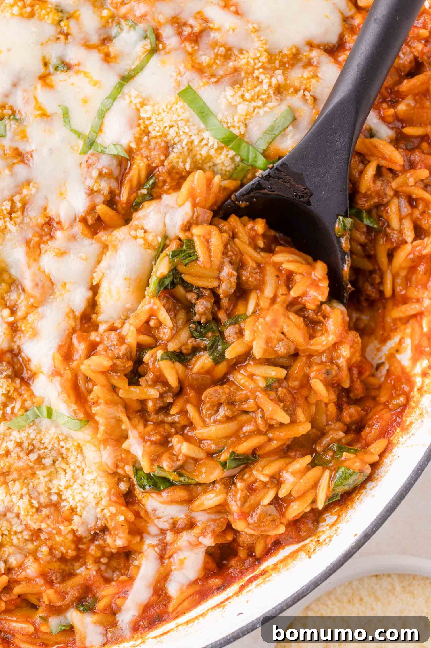 A close-up of a baked orzo pasta dish with ground meat, tomato sauce, melted cheese, and fresh basil, being served with a black spoon.