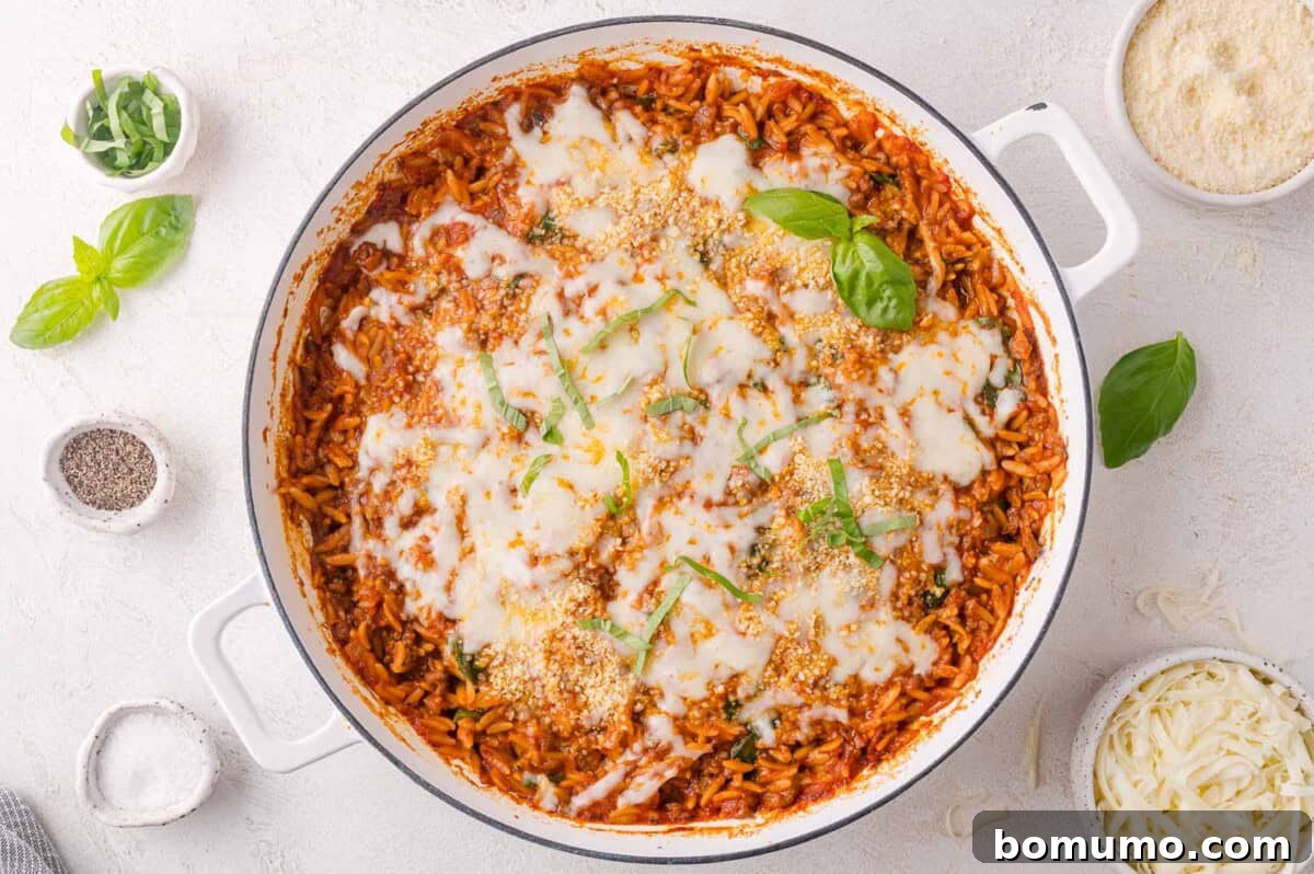 A white casserole dish filled with baked orzo pasta in tomato sauce, topped with melted cheese and garnished with fresh basil, surrounded by small bowls of cheese, pepper, and herbs.