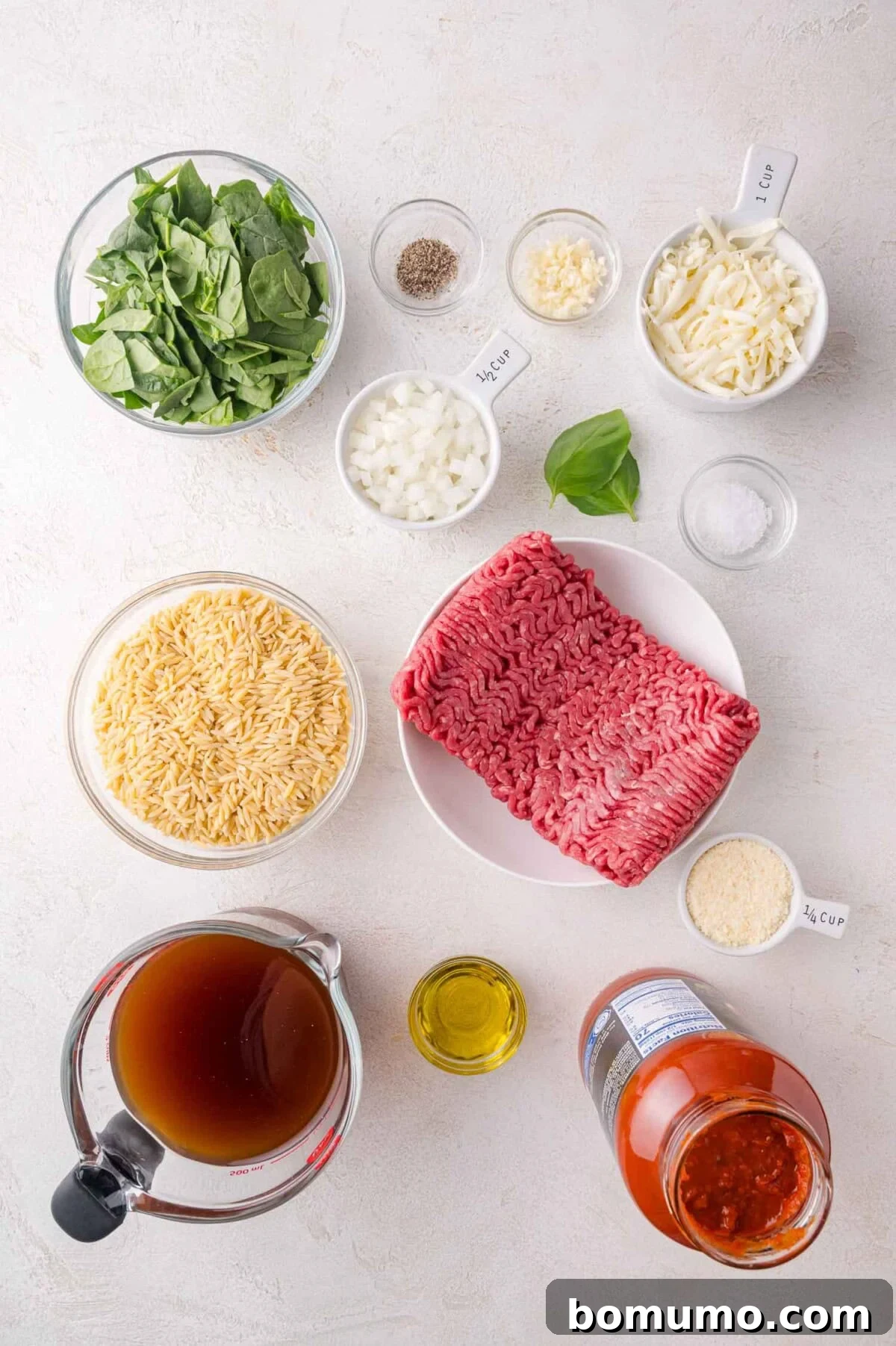 Ingredients for a pasta dish arranged on a white surface, including ground beef, orzo, spinach, cheese, onions, garlic, olive oil, broth, marinara sauce, and seasonings in bowls and measuring cups.