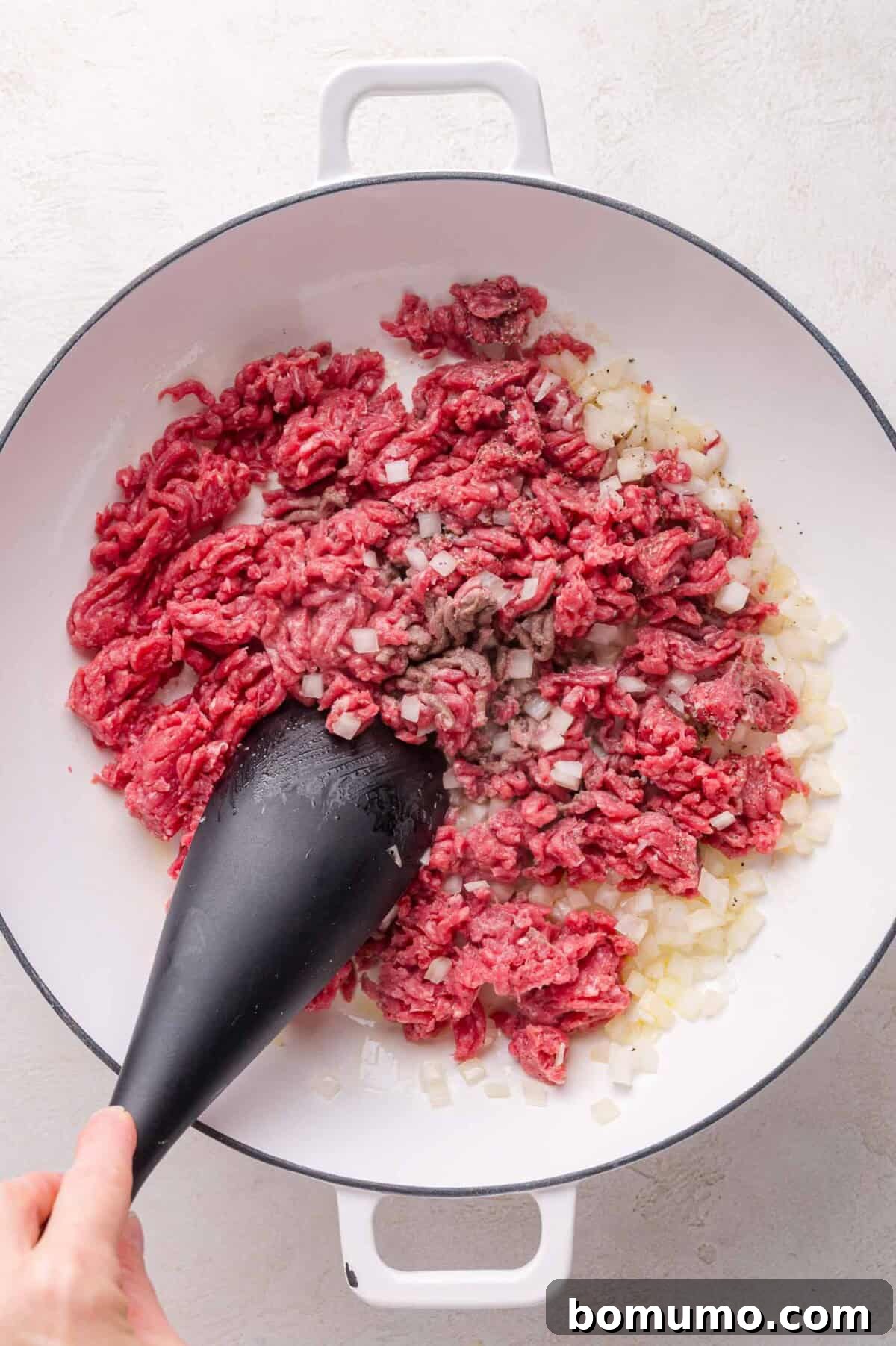 Ground beef and chopped onions being sautéed in a white pan, stirred with a black spatula.