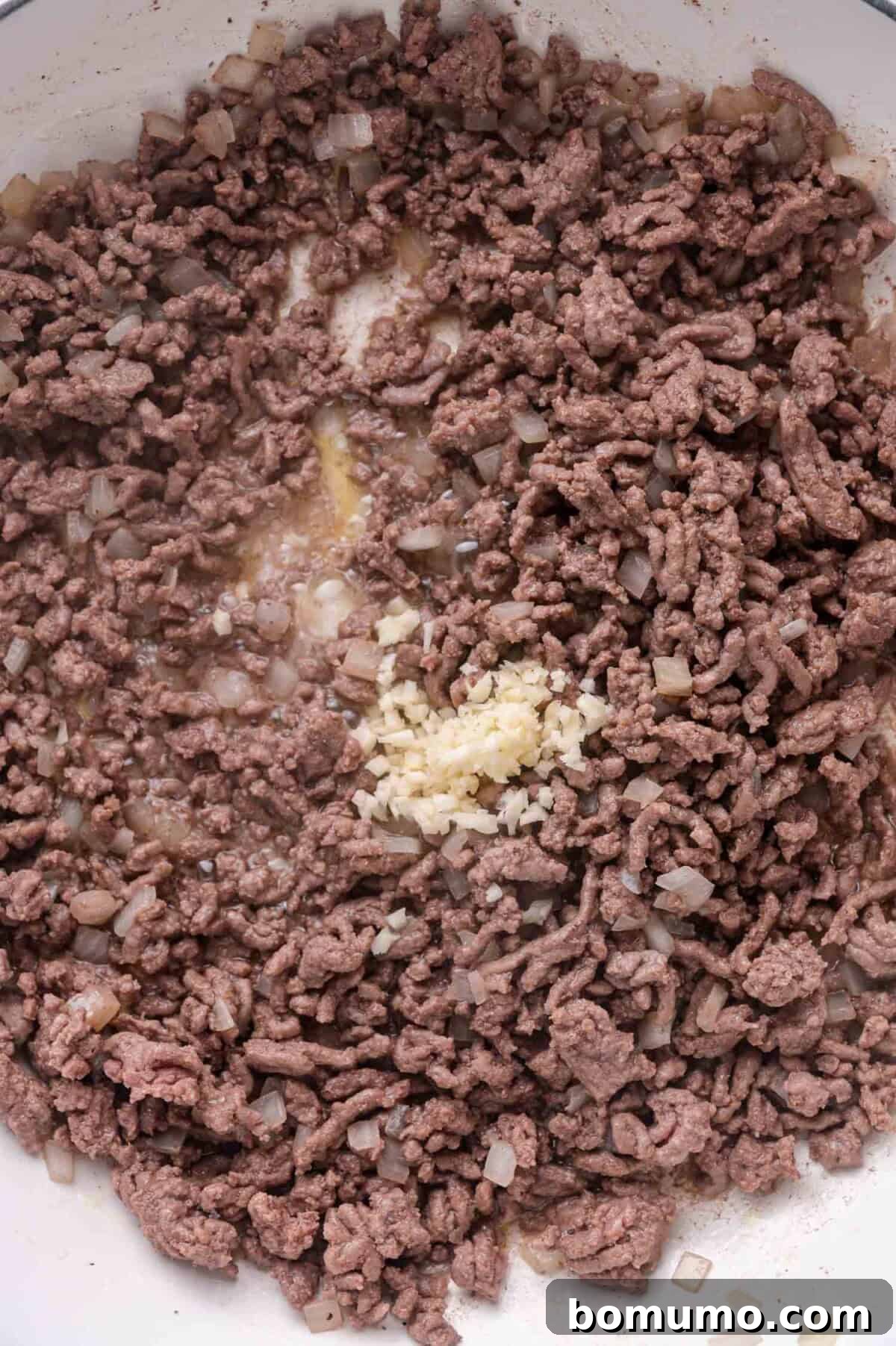 Cooked ground beef and diced onions in a pan with a small pile of minced garlic added to the center.
