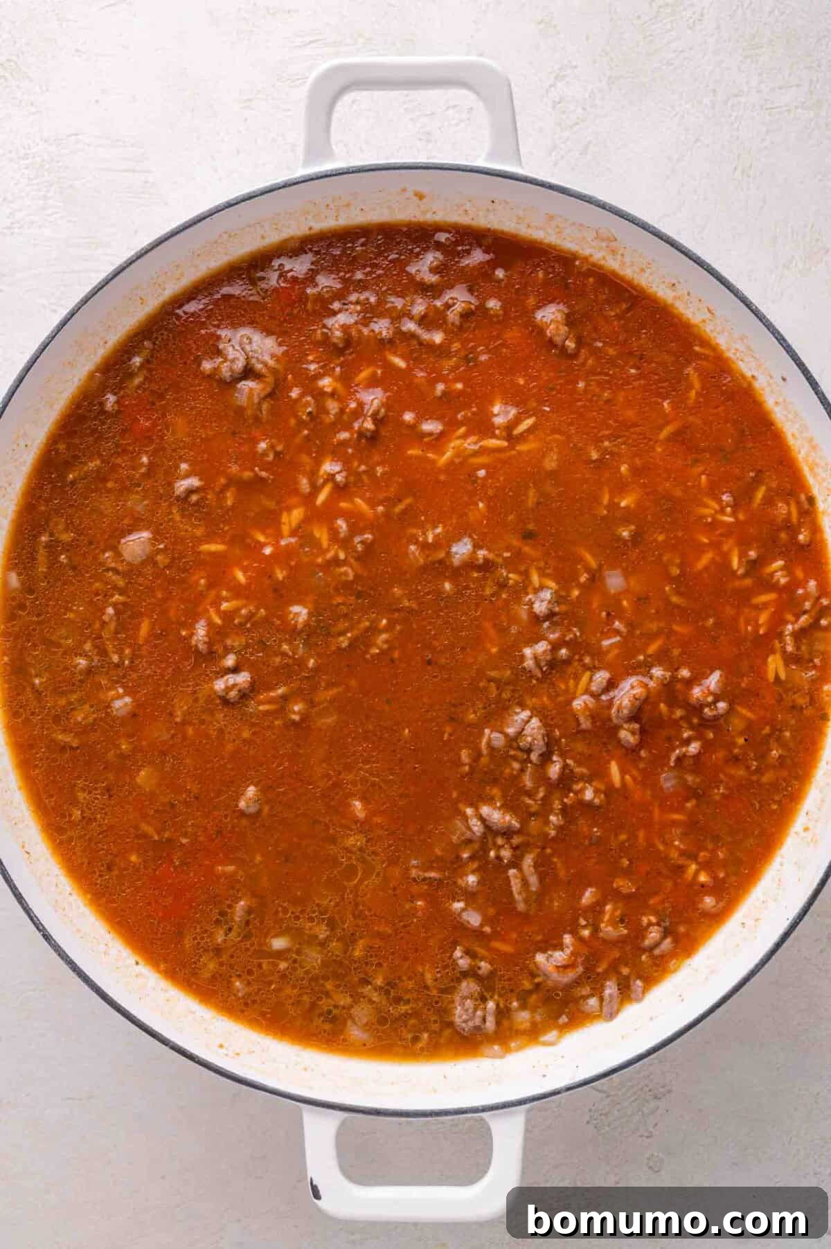 Large white pot filled with tomato-based mixture containing ground meat, orzo, and herbs, placed on a light-colored surface.