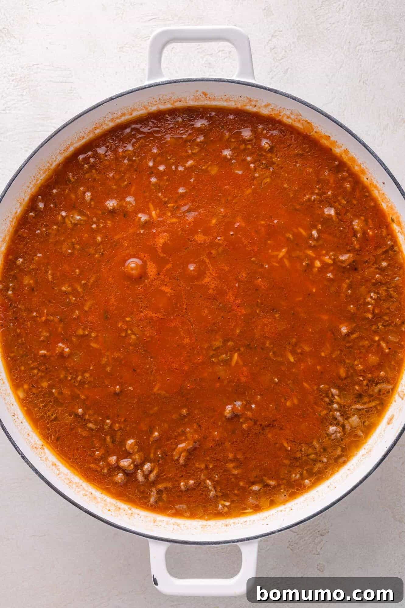 Large white pot filled with chunky red meat sauce, simmering, seen from above on a light surface.