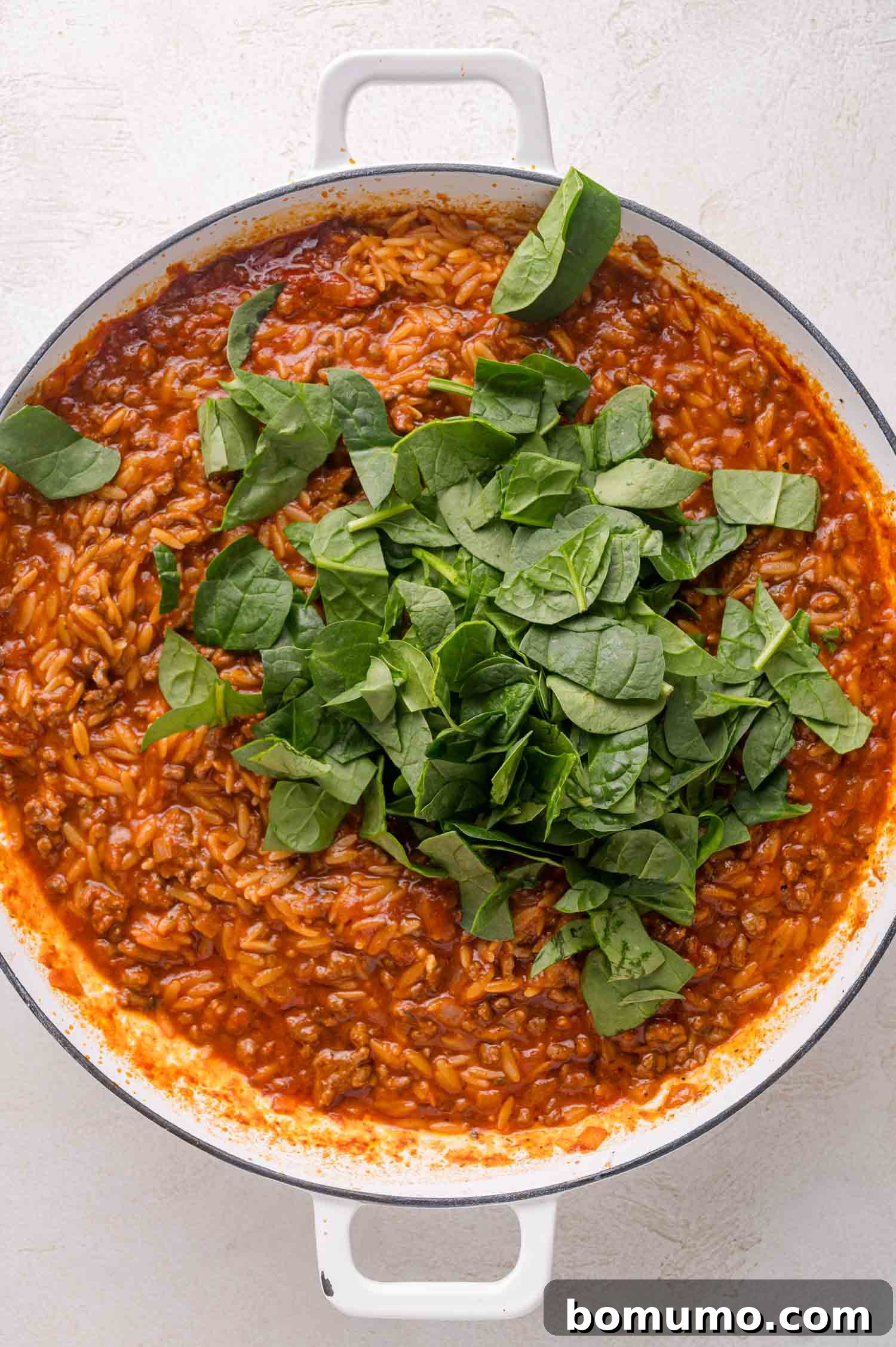 A white pot filled with tomato-based orzo pasta topped with fresh spinach leaves on a light surface.