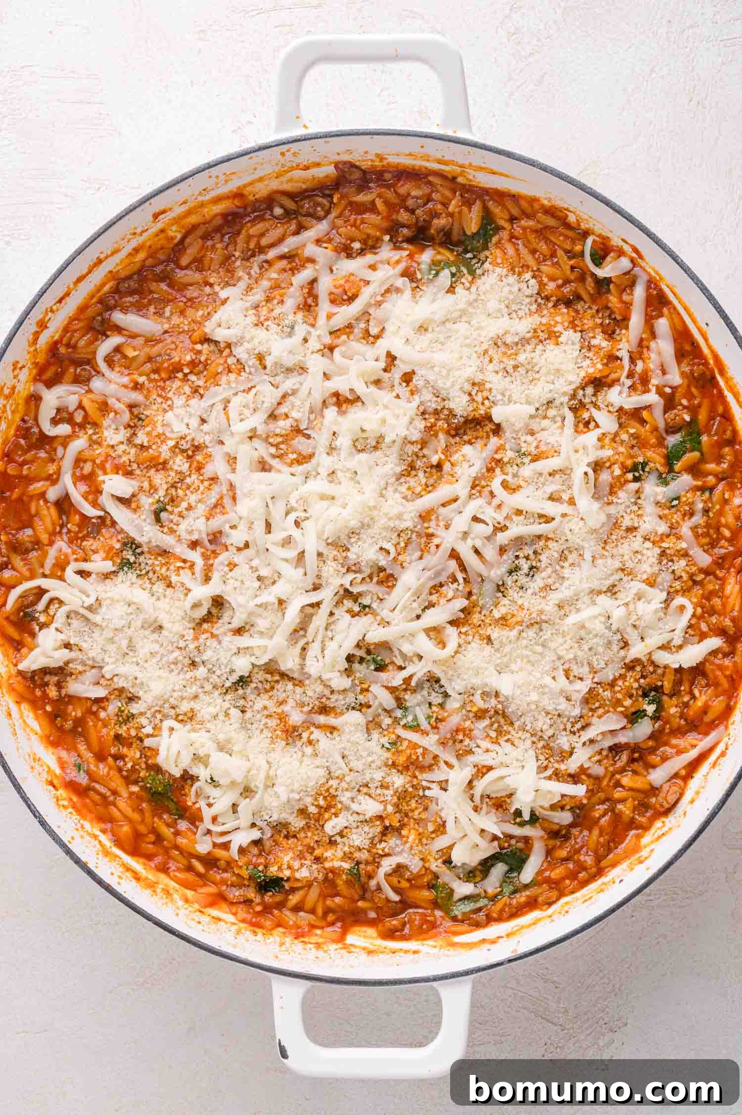 A white pot filled with tomato-based orzo pasta topped with shredded mozzarella and grated parmesan cheese.