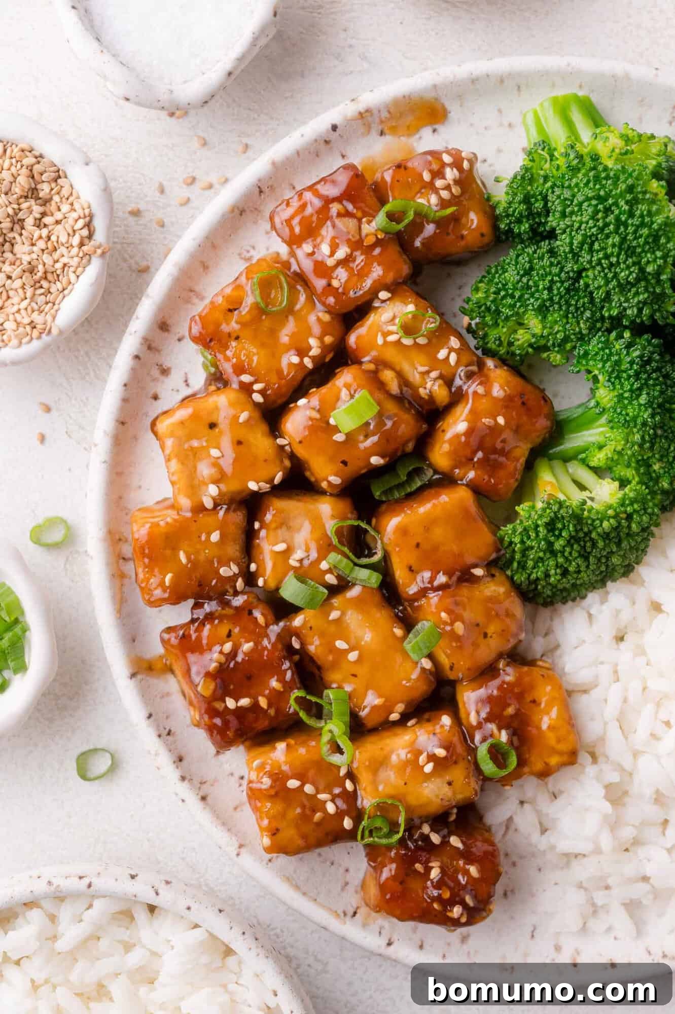 A plate with glazed teriyaki tofu cubes topped with sesame seeds and chopped green onions, served with steamed broccoli and white rice.
