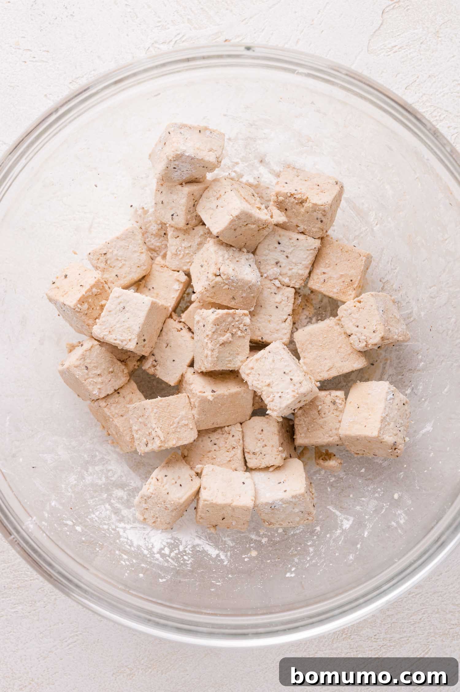 Cubed tofu coated in a light layer of seasoning and cornstarch sits in a clear glass mixing bowl on a white surface.