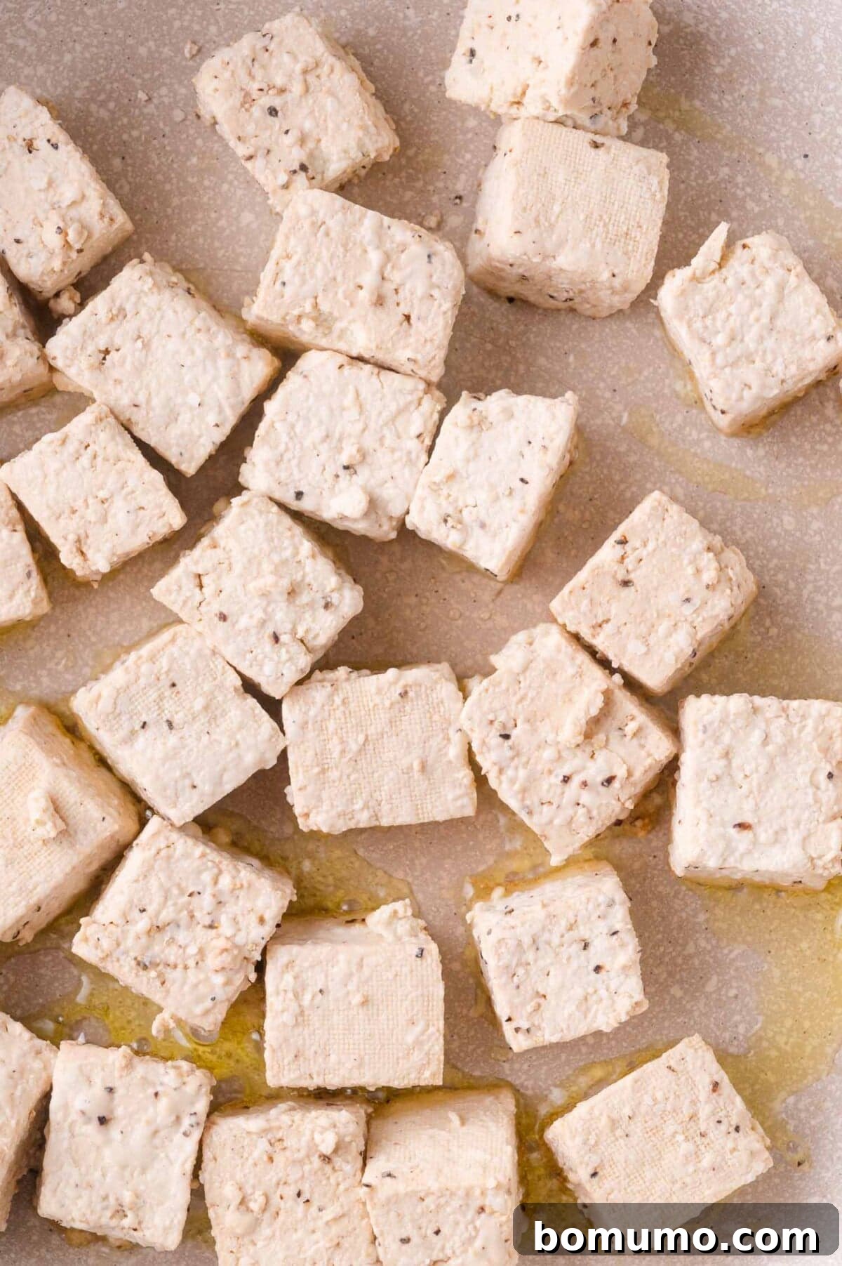 Cubed tofu pieces seasoned with pepper and scattered on a light-colored surface with a drizzle of oil.