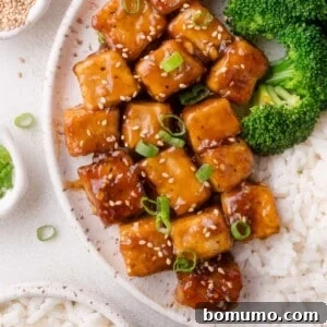 A plate with glazed teriyaki tofu cubes topped with sesame seeds and green onion, served with steamed broccoli and white rice.