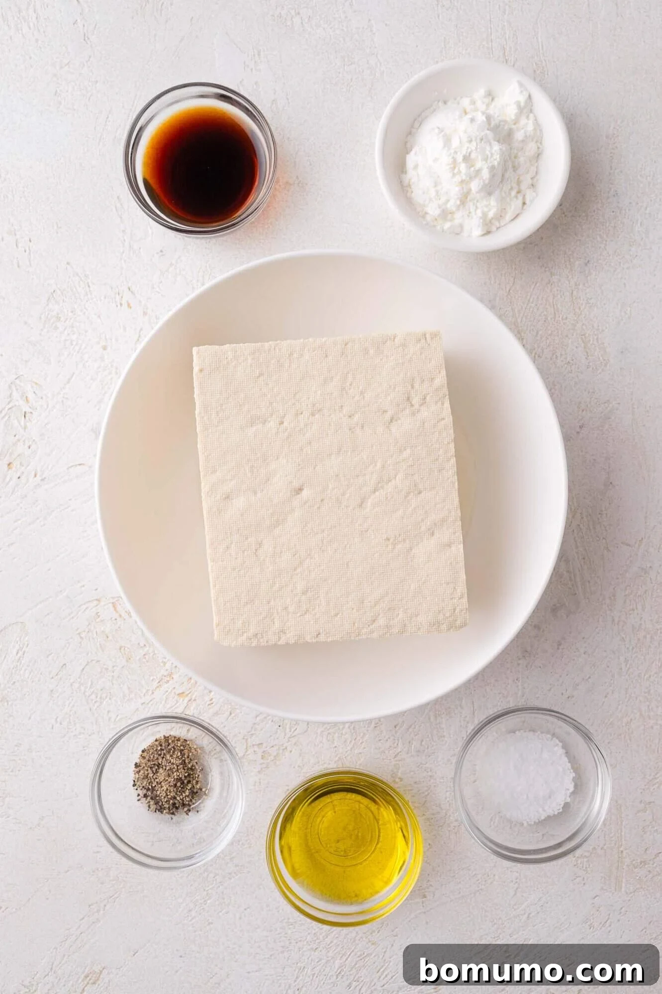 A block of tofu on a white plate surrounded by small bowls of soy sauce, cornstarch, ground pepper, olive oil, and salt.