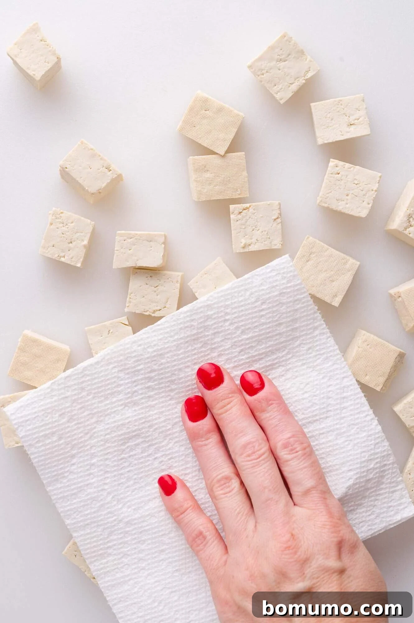 A hand with red nail polish presses a paper towel onto cubes of tofu on a white surface.