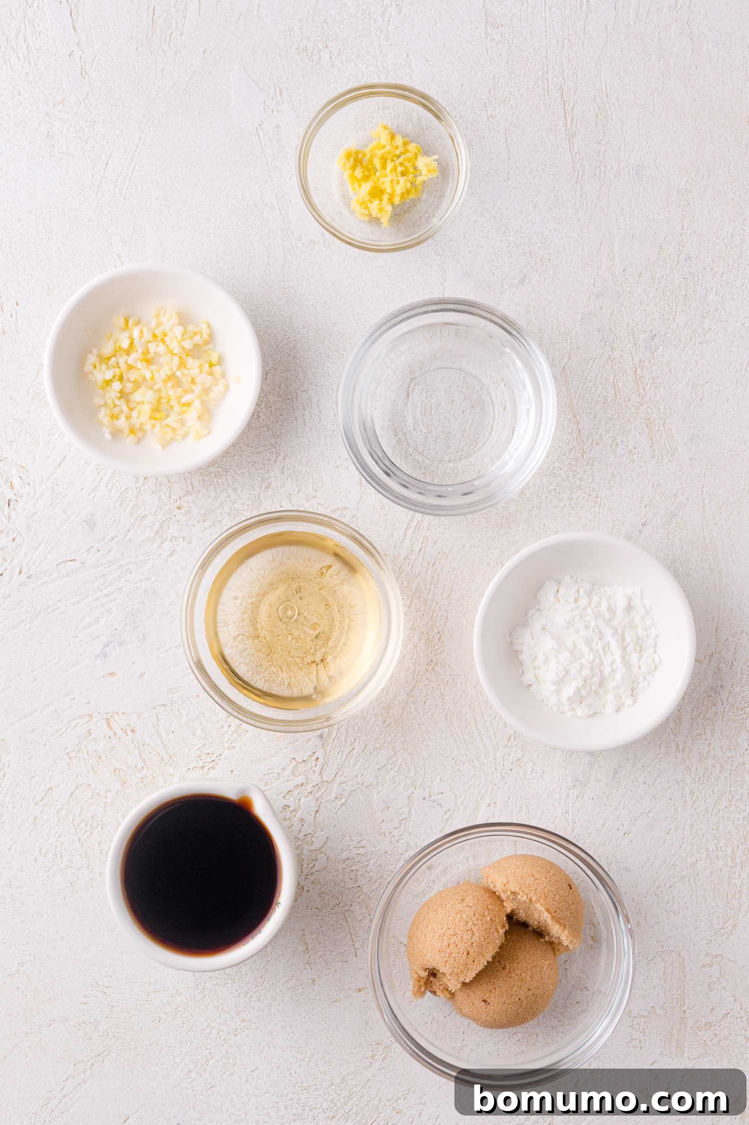 Seven small bowls containing minced garlic, minced ginger, water, rice vinegar, corn starch, soy sauce, and brown sugar arranged on a white surface.