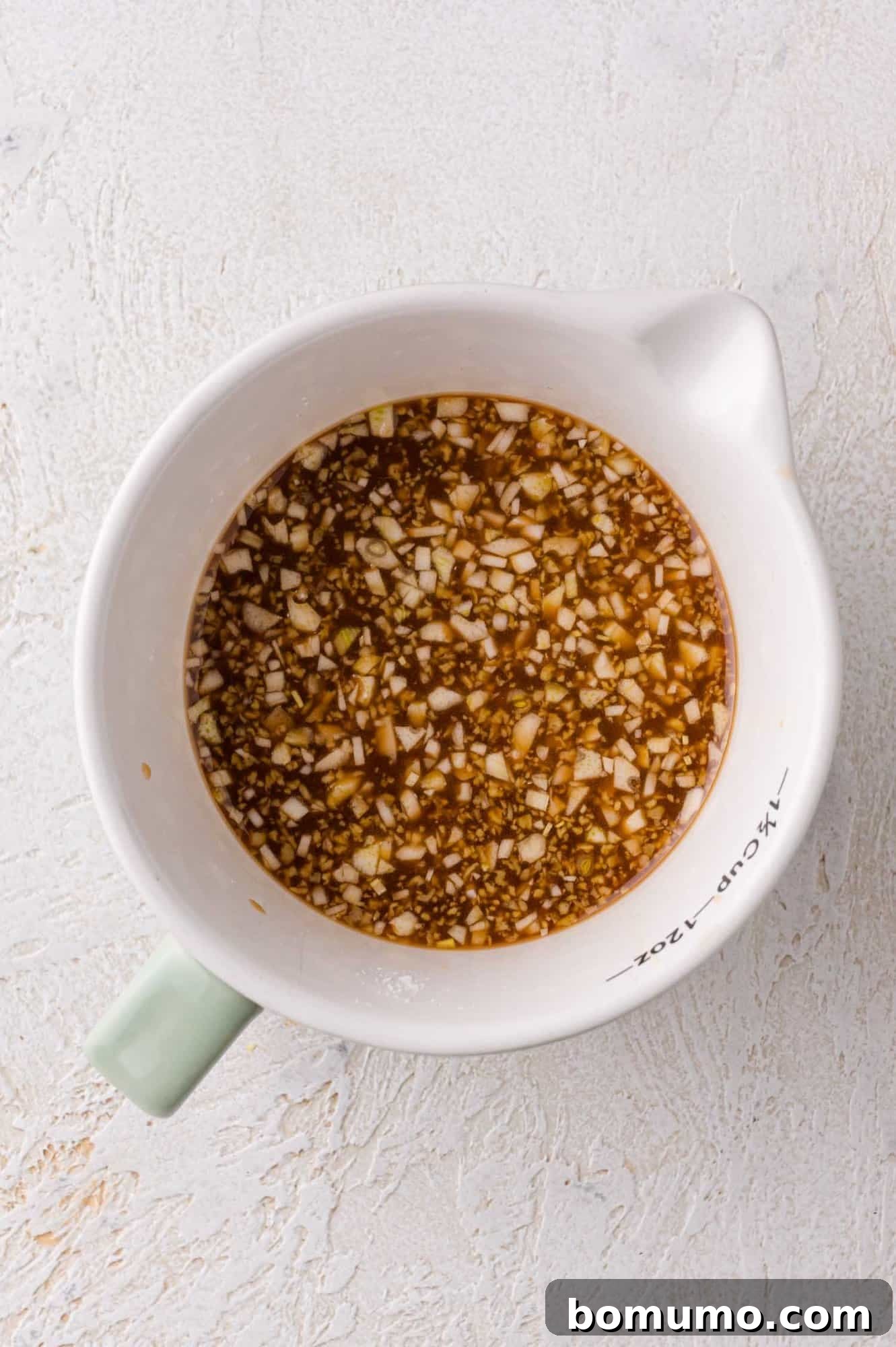 A white measuring cup contains a chopped garlic and soy sauce mixture on a light textured surface.