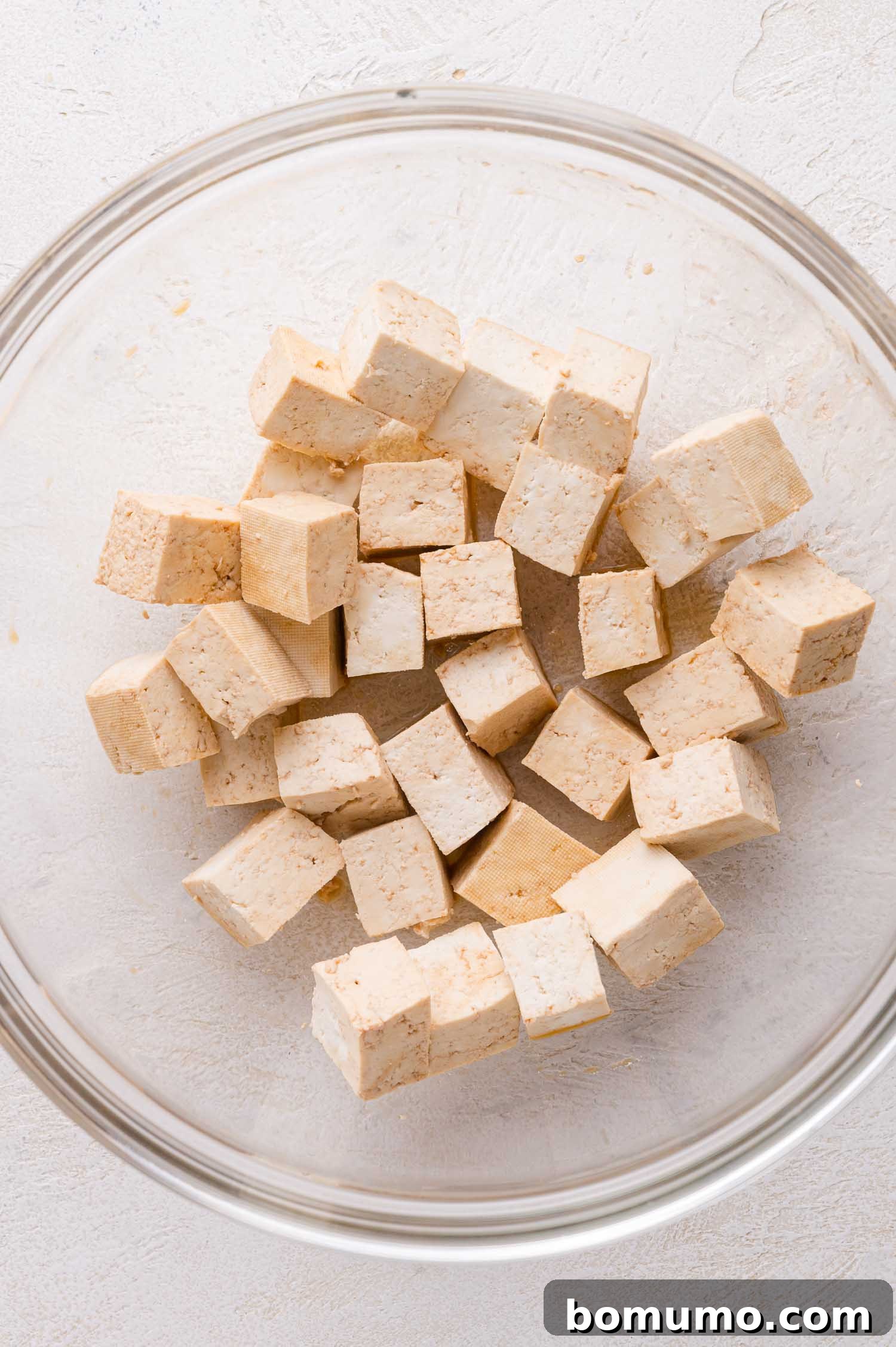 A clear glass bowl containing several cubes of plain tofu on a light-colored surface.