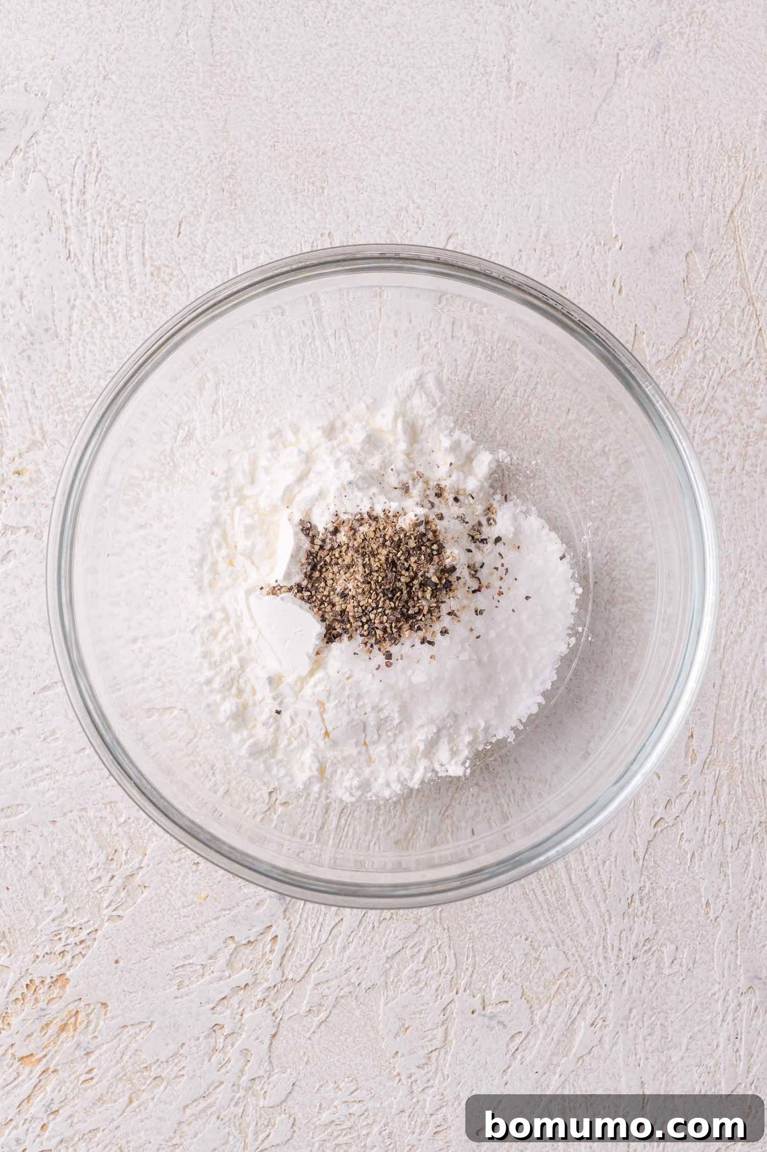 A glass bowl containing a mixture of cornstarch and a small amount of ground black pepper.