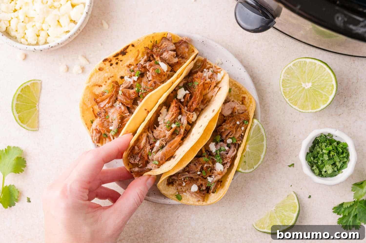 Hand holding a taco with shredded carnitas, cheese, and herbs; another taco on the plate; surrounded by lime wedges, cilantro, cheese, and a slow cooker.