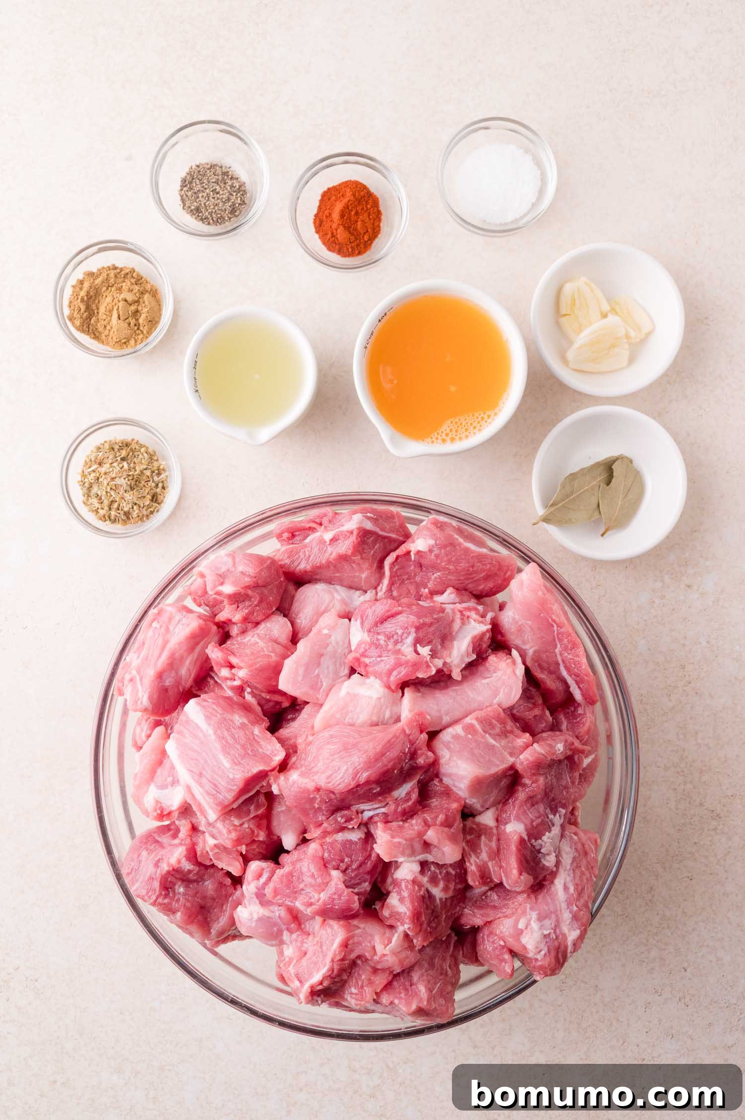 A glass bowl filled with raw pork chunks is surrounded by small bowls of spices, herbs, garlic cloves, vinegar, salt, pepper, and bay leaves on a light surface.