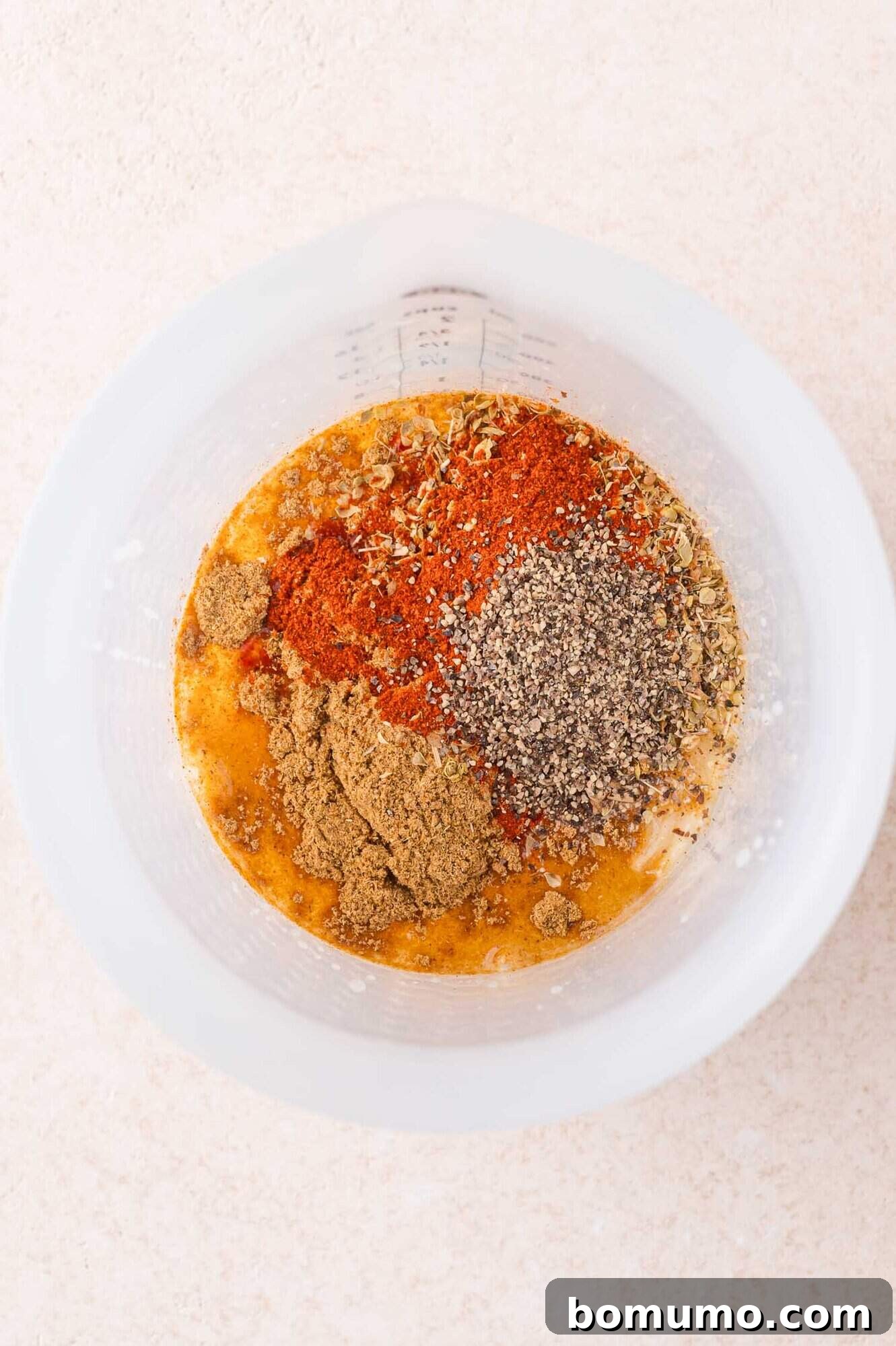 A mixing bowl containing ground spices, black pepper, and liquid ingredients arranged in separate piles before mixing.