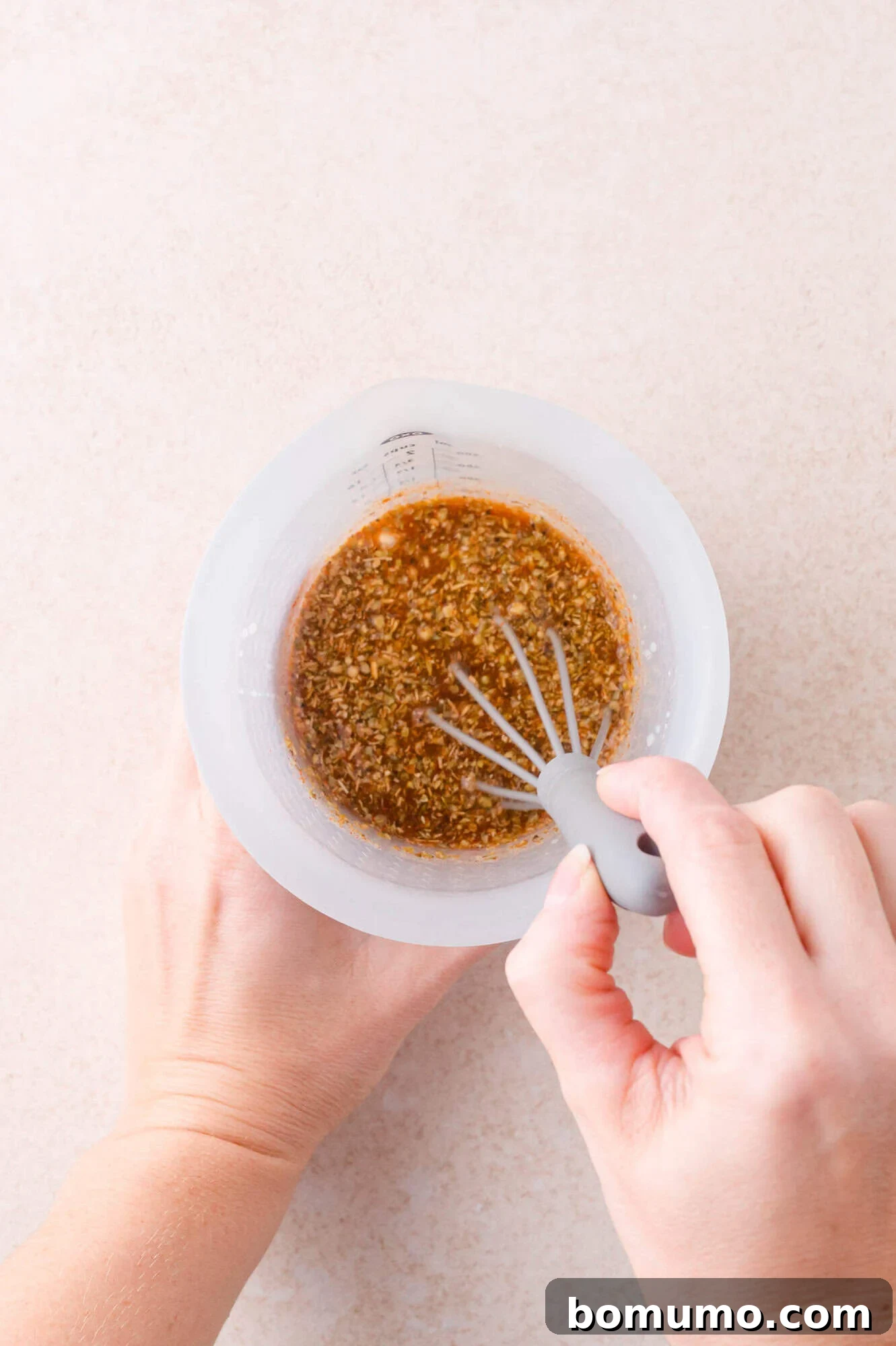 A person uses a small whisk to mix a marinade or sauce with visible minced ingredients in a clear measuring cup on a light surface.