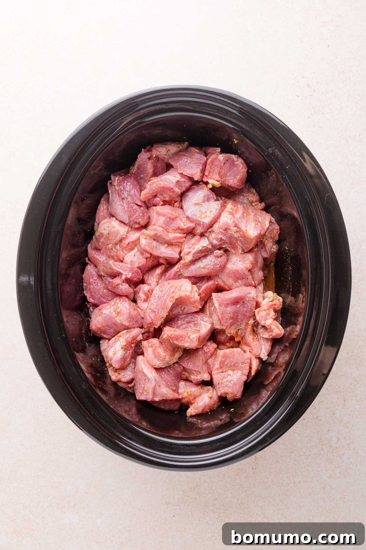 Chunks of raw, seasoned pork are placed inside a black oval slow cooker on a light-colored surface.