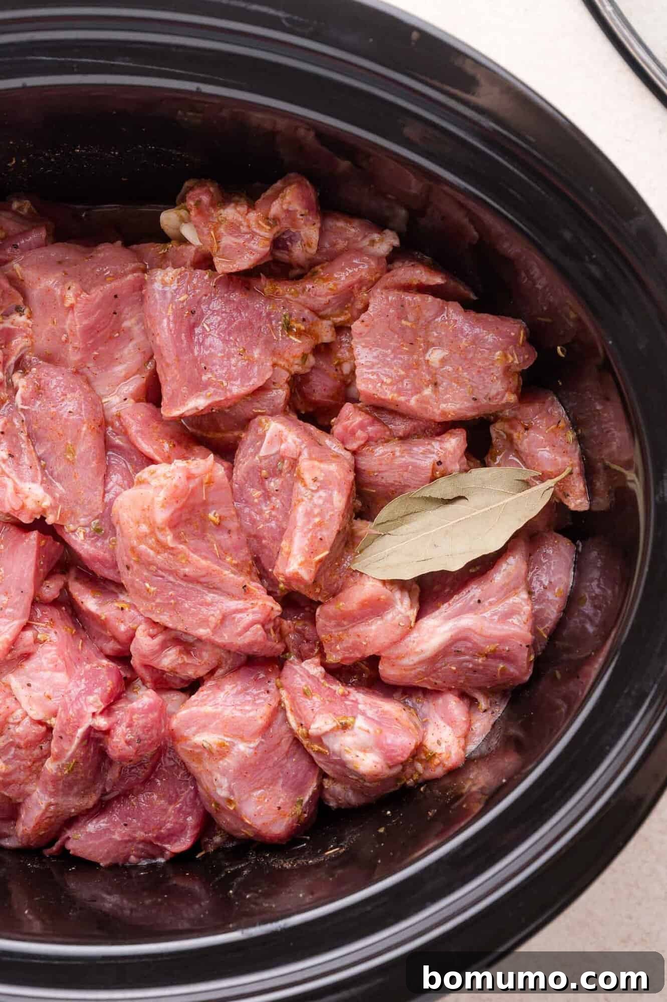 Chunks of raw, seasoned pork and a bay leaf in a black slow cooker, ready to be cooked.
