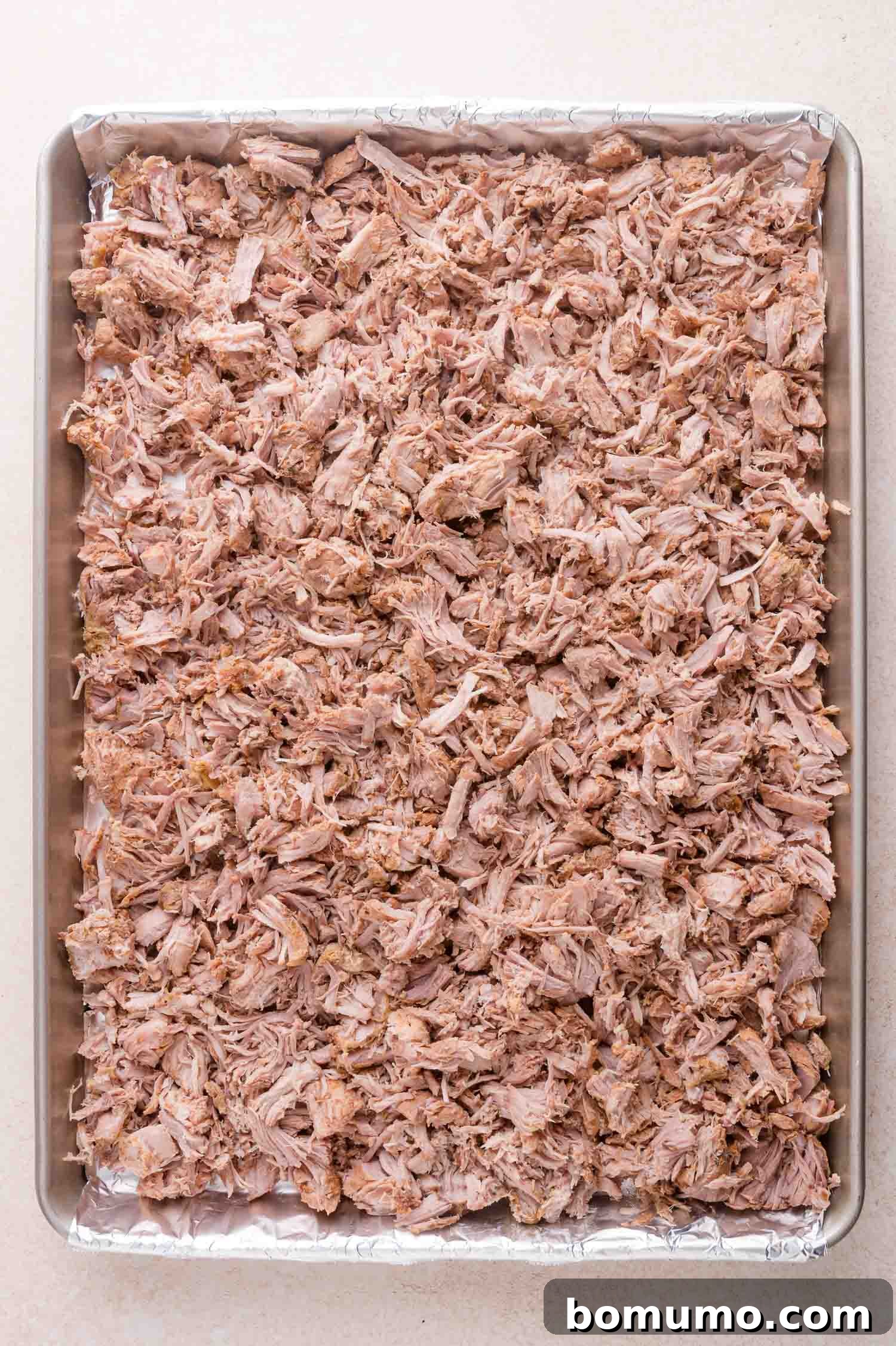 A baking sheet lined with foil is filled with a large amount of shredded cooked pork spread evenly across the surface.