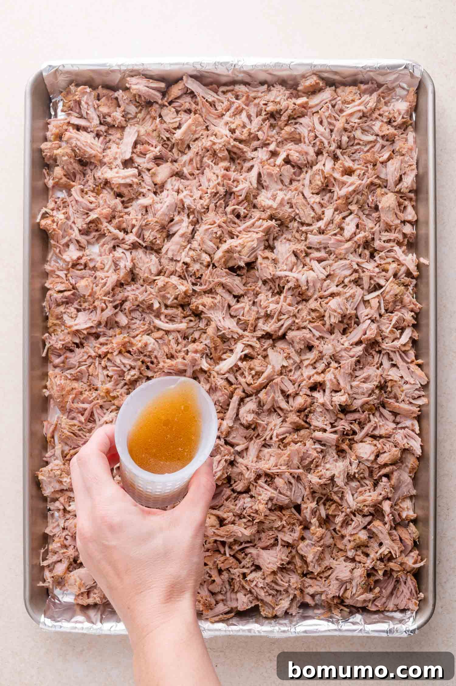 A hand pours broth from a cup over a baking sheet covered with shredded cooked pork.