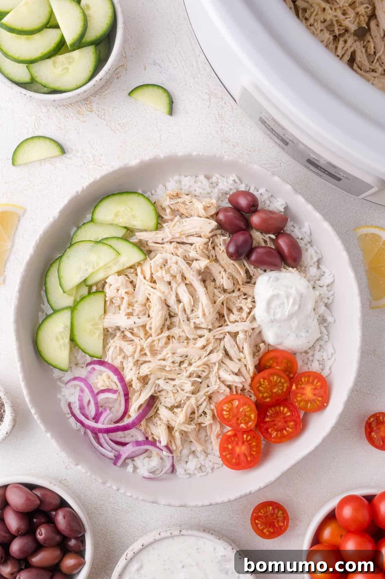 A vibrant Greek chicken bowl with shredded chicken, fresh cucumbers, cherry tomatoes, red onions, Kalamata olives, and a dollop of creamy tzatziki sauce, served over fluffy rice, with a slow cooker and extra ingredients in the background.