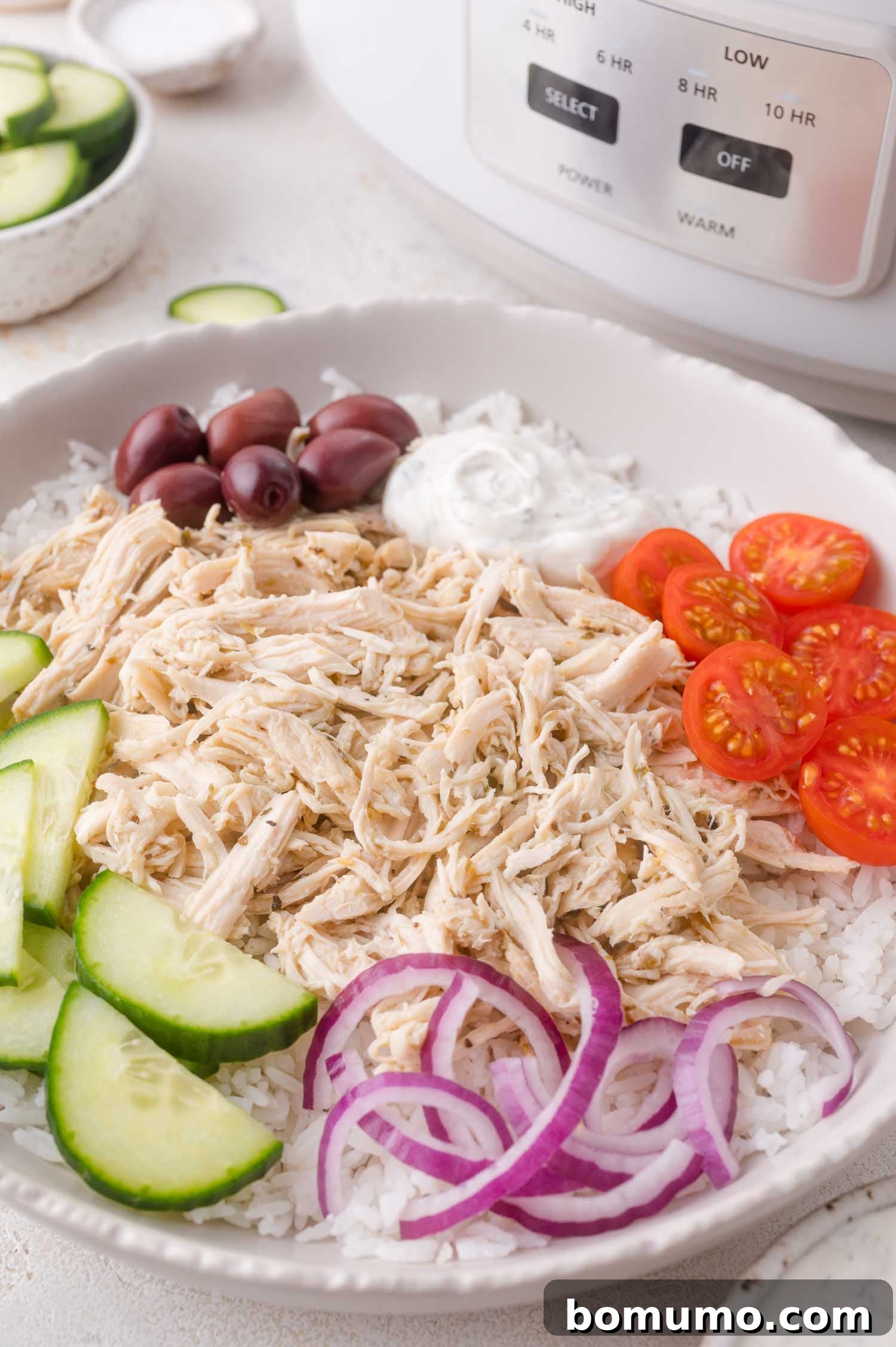 A beautifully presented bowl of shredded Greek chicken served over white rice, garnished with vibrant sliced cucumbers, cherry tomatoes, red onions, briny Kalamata olives, and a dollop of creamy white sauce.