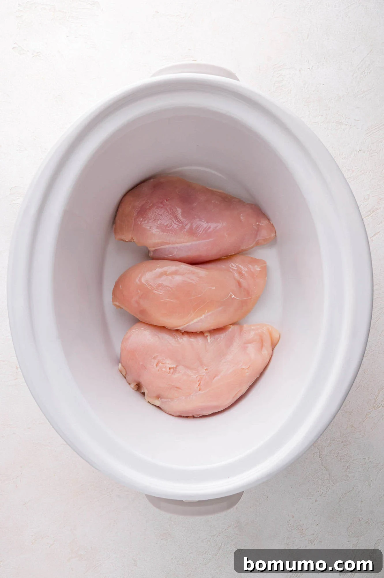 Three raw chicken breasts are neatly placed in a single layer inside a white slow cooker, prepared for cooking.