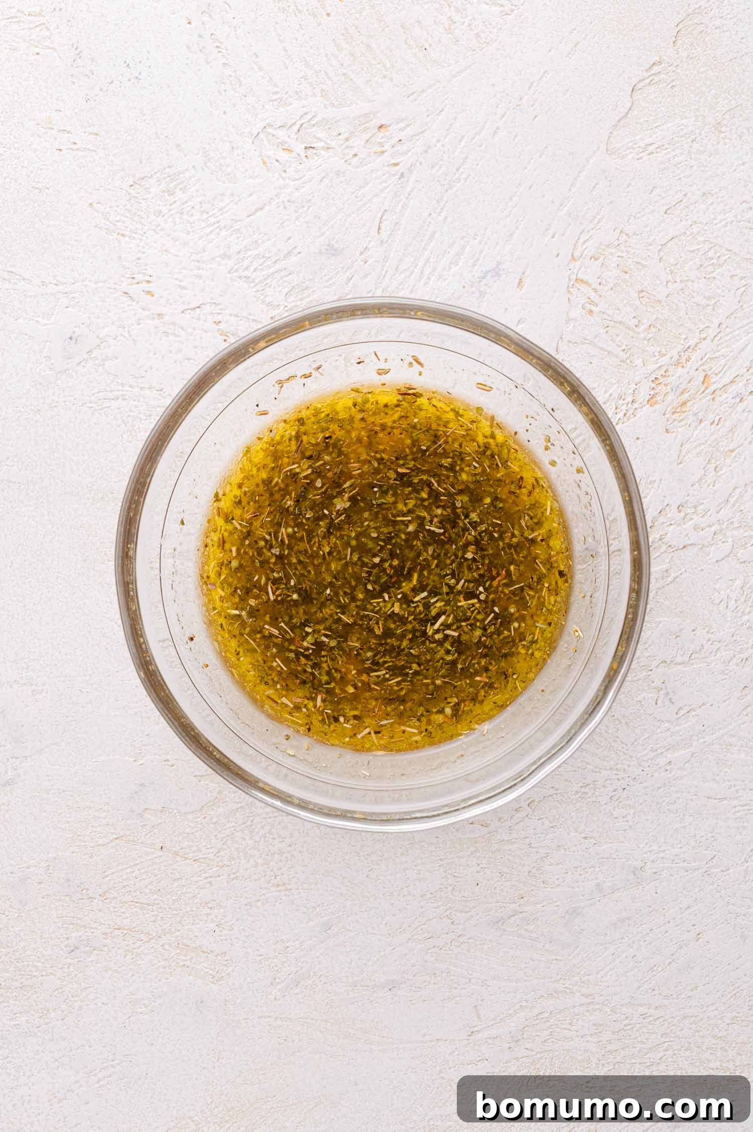 A clear glass bowl contains a perfectly emulsified mixture of olive oil, lemon juice, red wine vinegar, garlic, oregano, and other seasonings, ready to be poured over chicken.