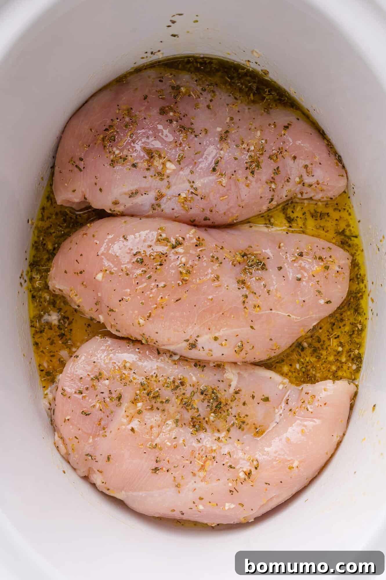 Raw chicken breasts, beautifully coated in the Greek marinade, rest in a white slow cooker, ready for the slow cooking process to begin.
