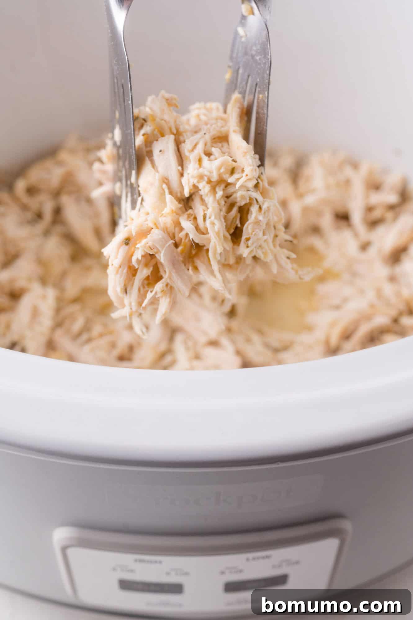 A pair of forks expertly pulling apart shredded chicken directly within the slow cooker, allowing it to reabsorb all the delicious cooking juices.