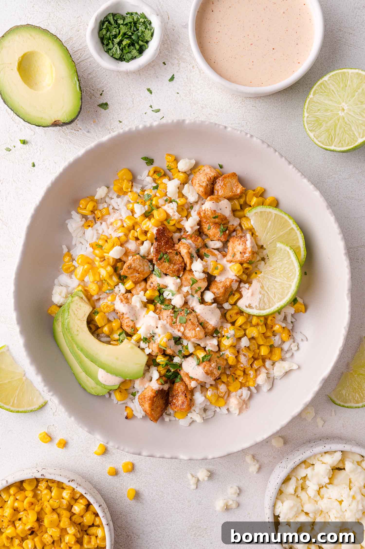 A vibrant bowl of rice topped with seasoned chicken, golden charred corn, fresh avocado slices, lime wedges, and crumbled cotija cheese, accompanied by small side bowls of cilantro, sauce, and additional corn.