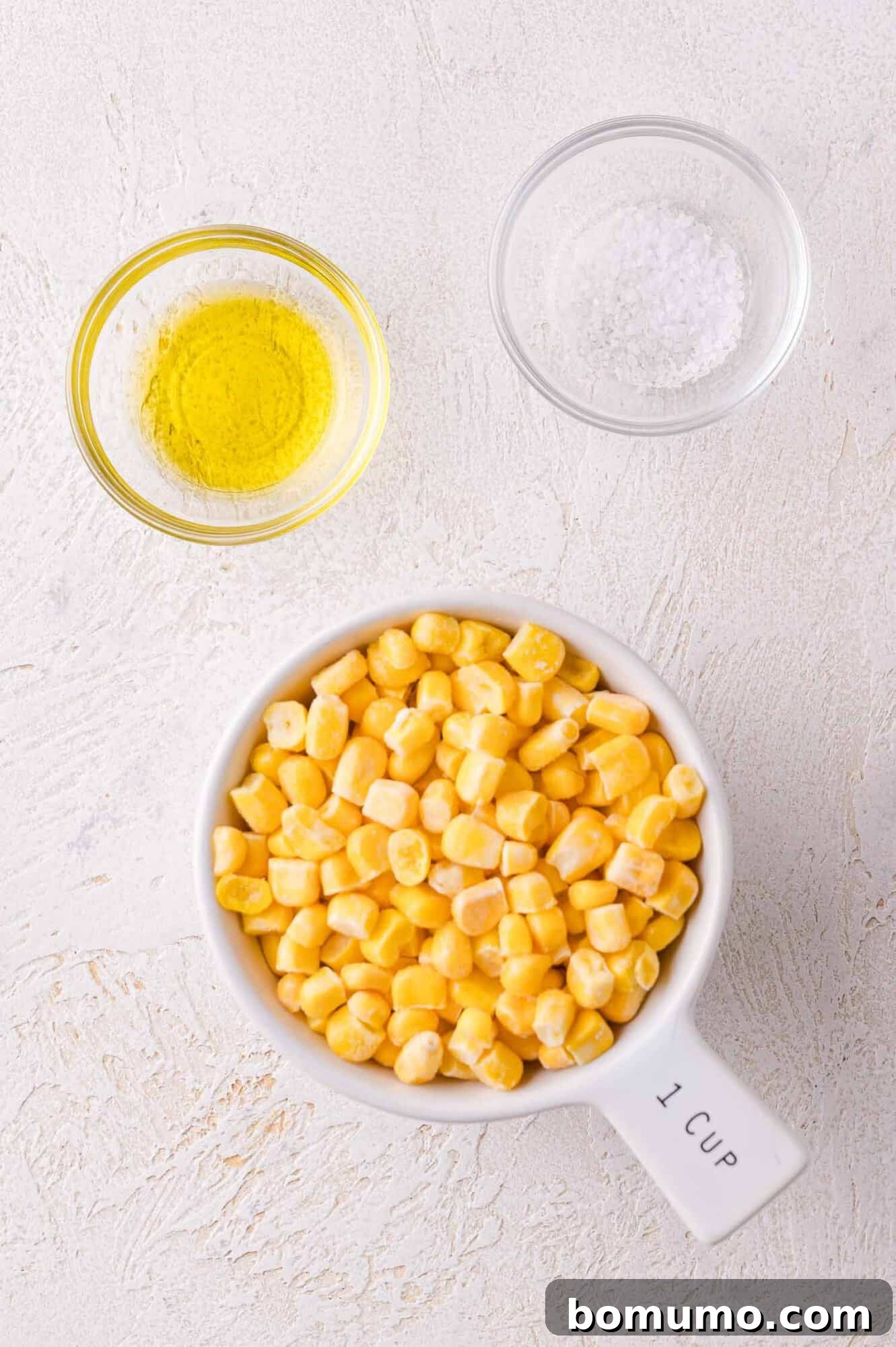 A clear measuring cup filled with bright yellow corn kernels sits alongside small bowls of olive oil and salt, ready for the next cooking step.