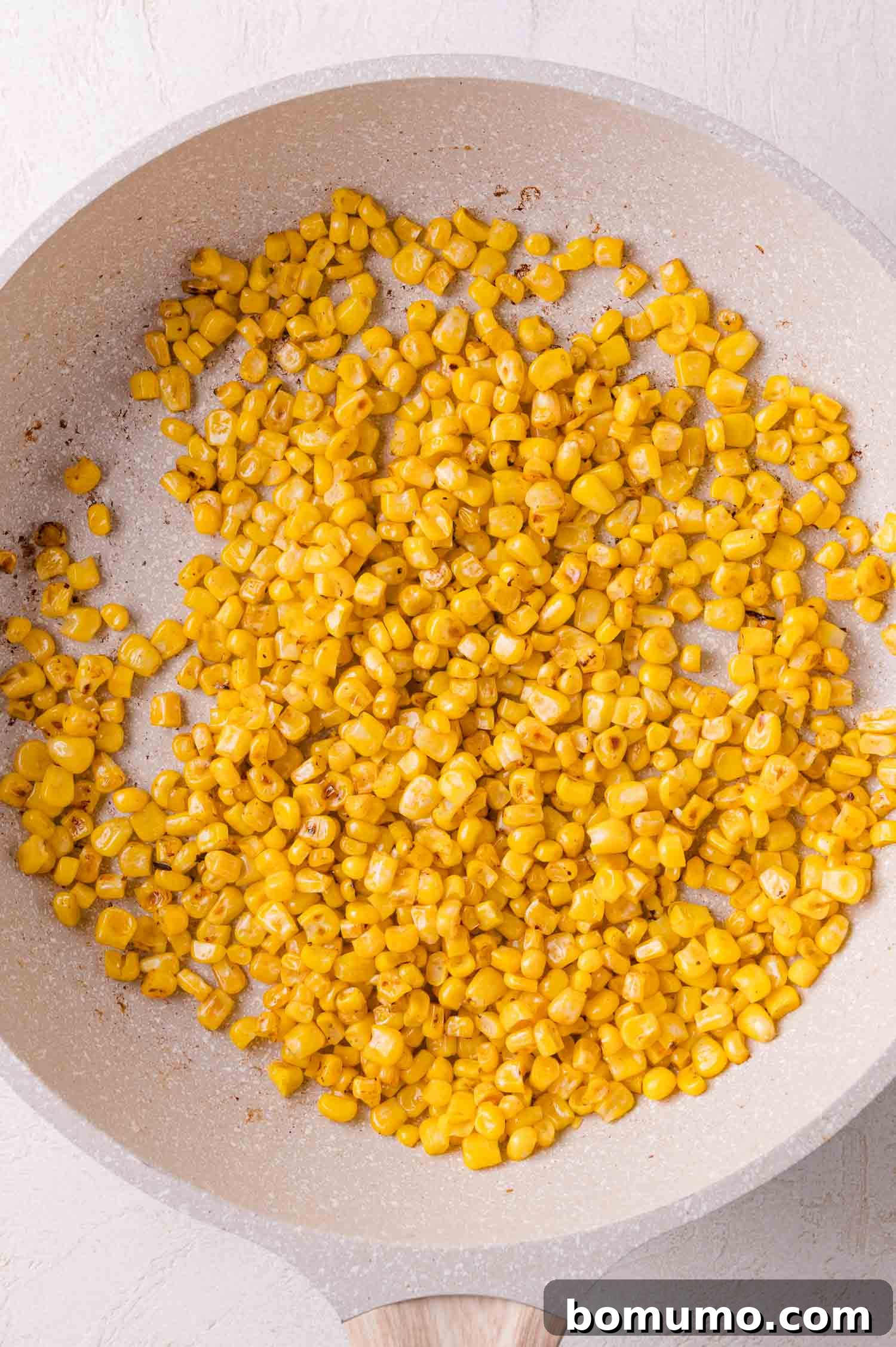 Vibrant corn kernels are being sautéed in a light-colored frying pan on a clean white surface, beginning to char and develop their sweet, smoky flavor.
