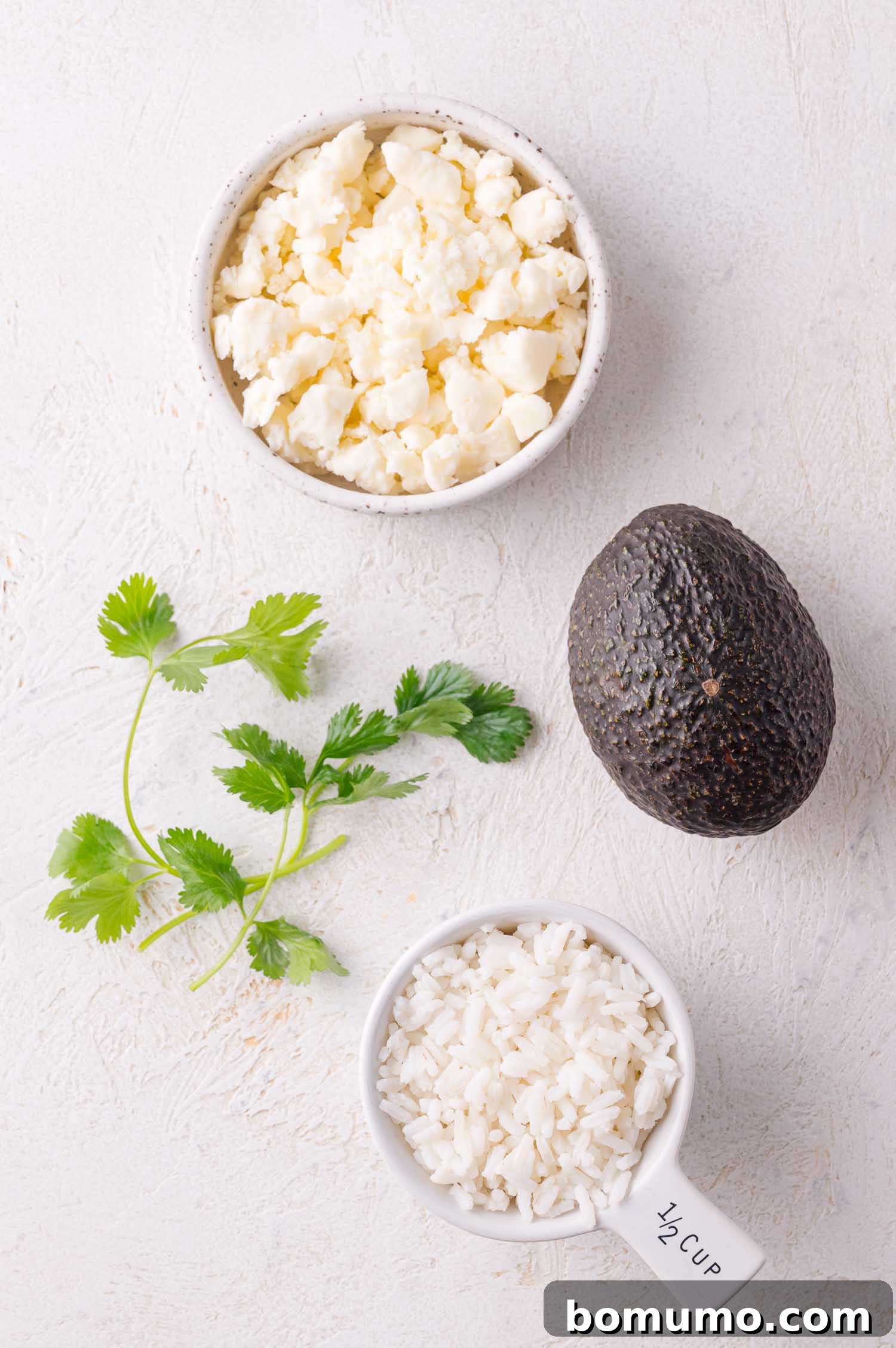 A collection of fresh ingredients for bowl assembly: a bowl of crumbled cotija cheese, a whole ripe avocado, a vibrant sprig of cilantro, and a measuring cup brimming with cooked rice, all arranged on a white surface.