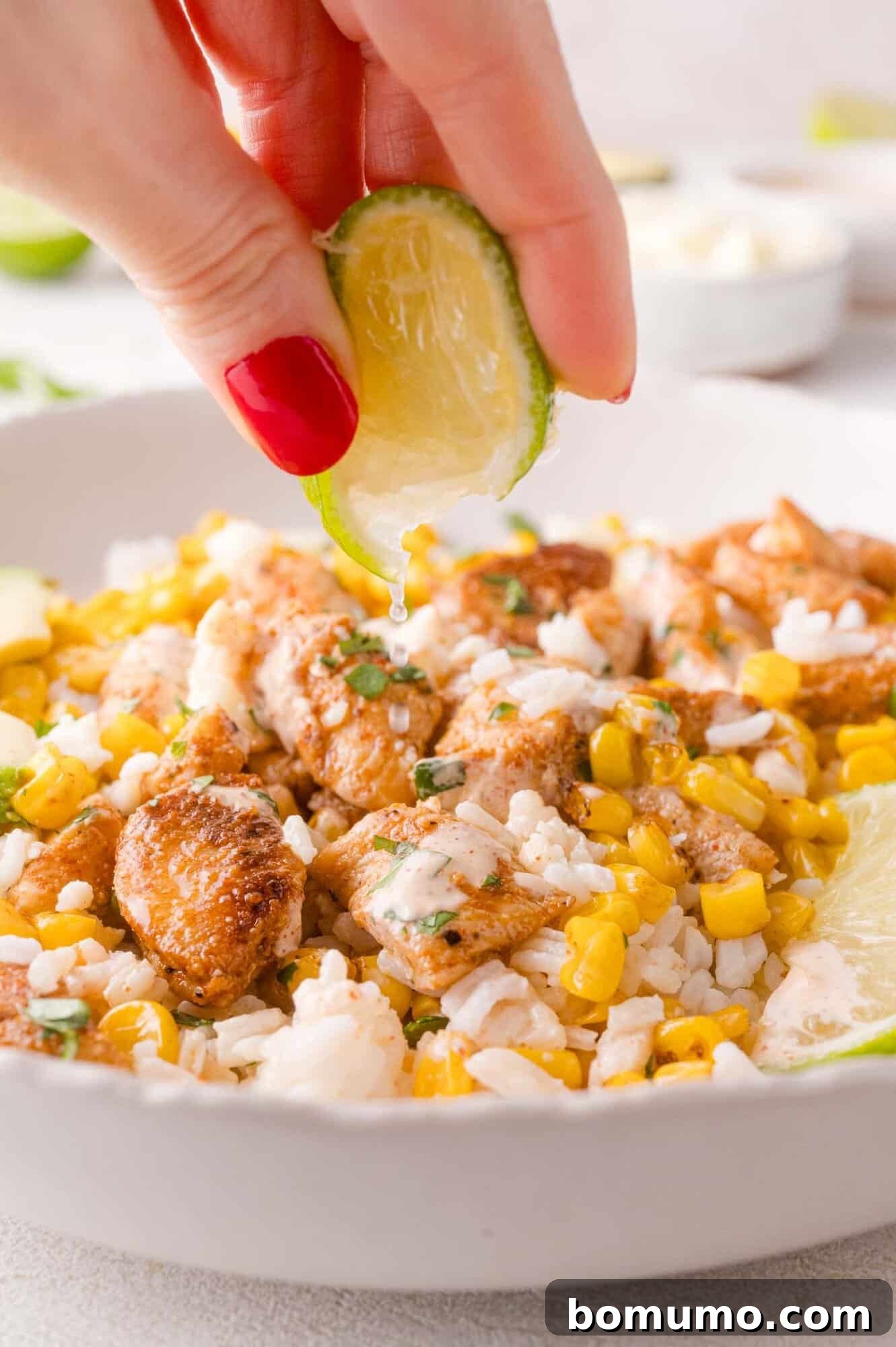 A hand expertly squeezes a fresh lime wedge over a meticulously prepared bowl, enriching the rice, corn, and seasoned chicken pieces with a vibrant burst of citrus.
