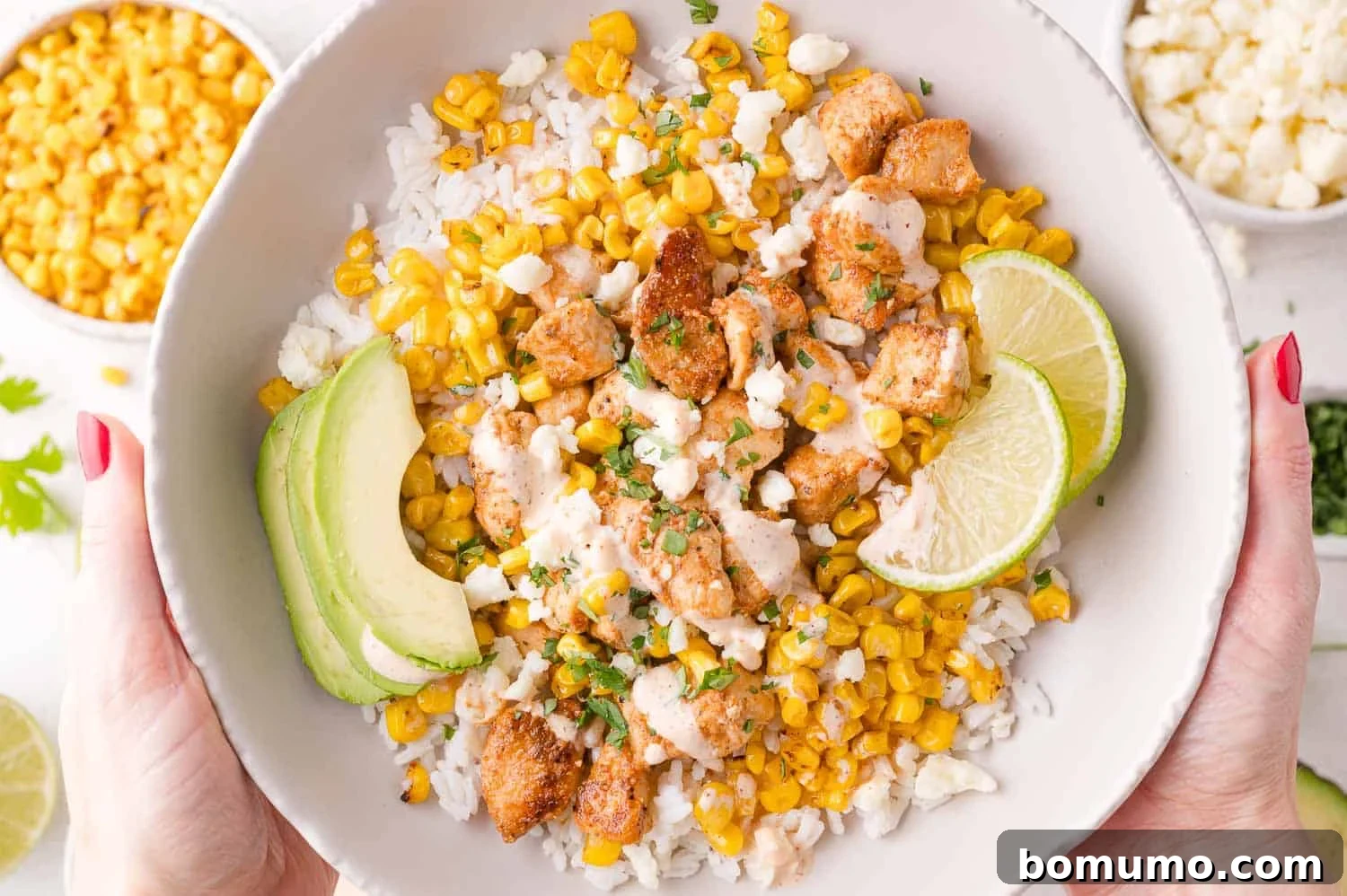 A vibrant bowl of rice topped with perfectly grilled chicken, golden charred corn, crumbled cheese, fresh avocado slices, and bright lime wedges, held invitingly in two hands, ready to be enjoyed.