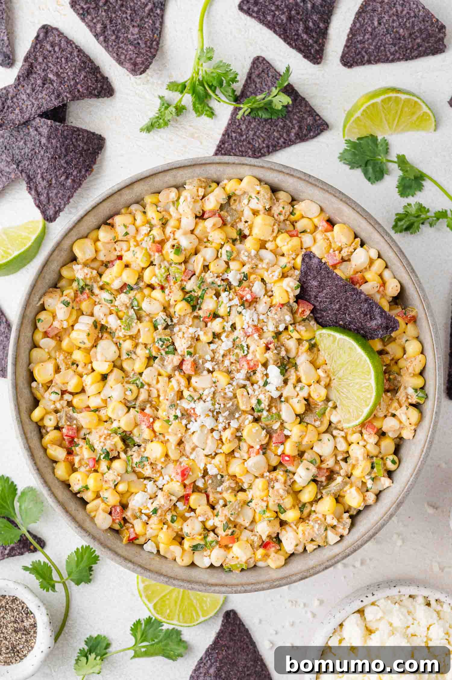 A bowl of Mexican street corn dip garnished with vibrant lime slices and crumbled cotija cheese, surrounded by crispy blue tortilla chips and fresh cilantro on a pristine white surface.