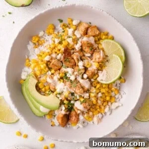A bowl of rice topped with grilled corn, seasoned chicken pieces, avocado slices, lime wedges, crumbled cheese, and a light drizzle of sauce.