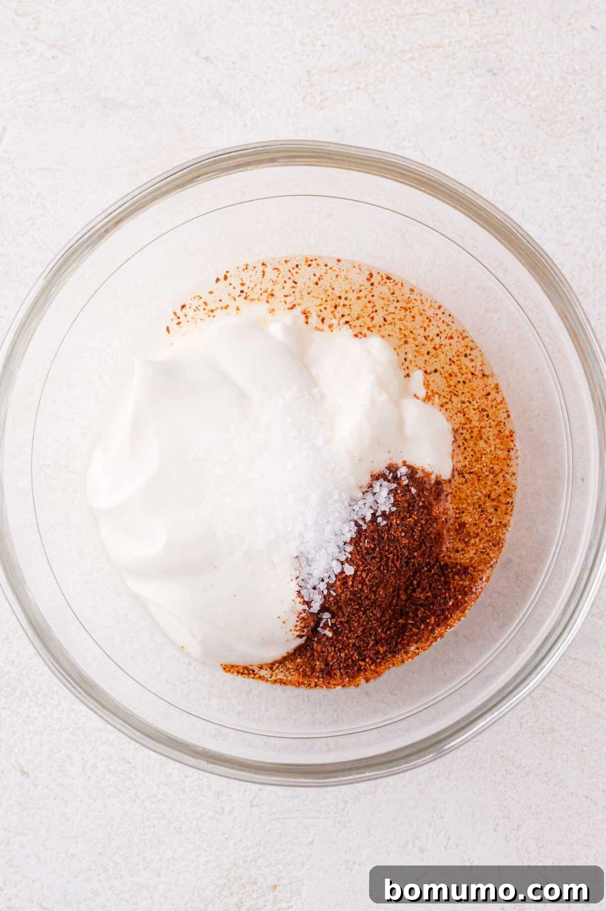 A glass bowl holds sour cream, chili powder, and salt, before whisking, capturing the initial blend of key sauce ingredients.