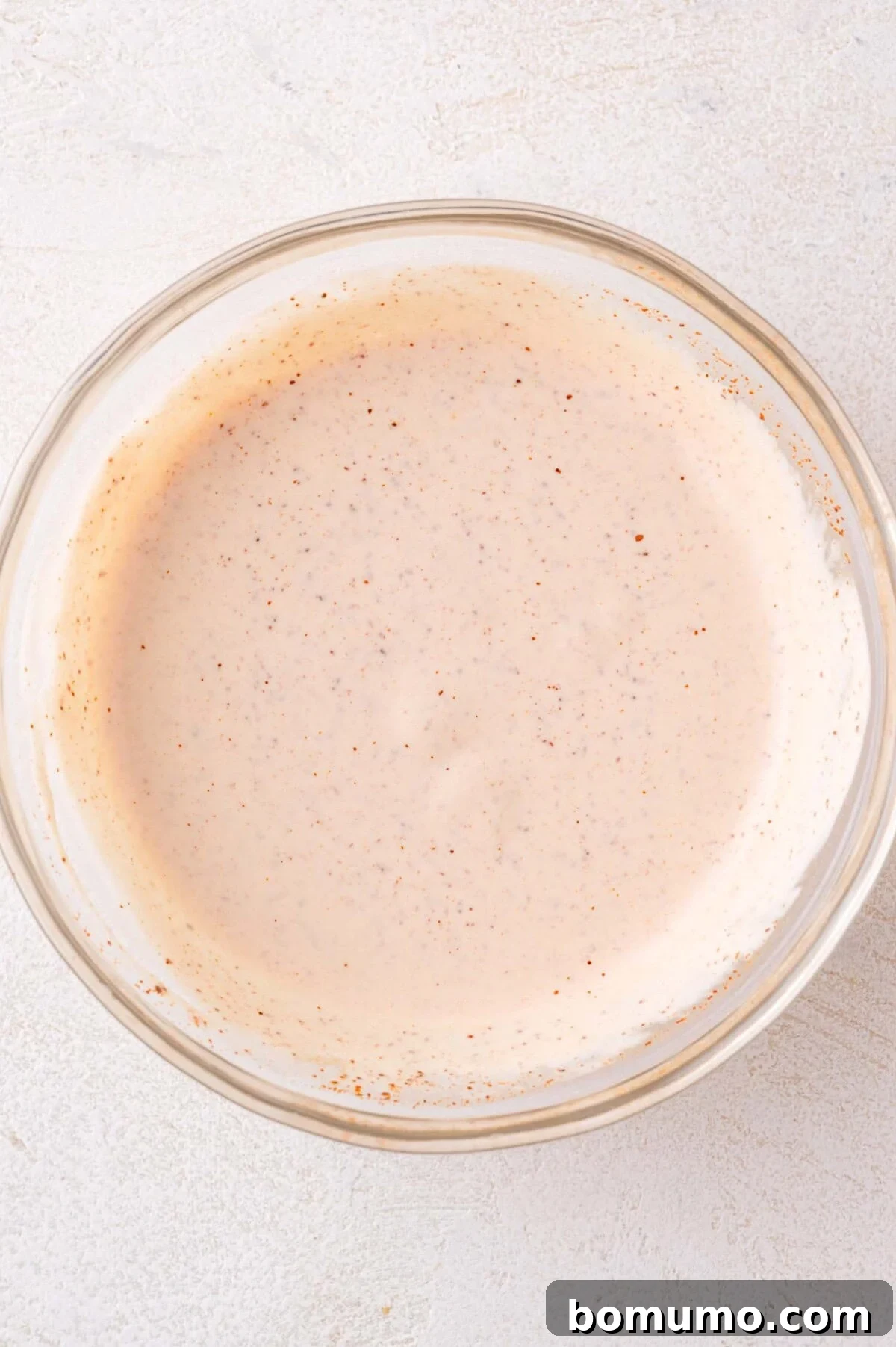 A clear glass bowl showcases the creamy, light orange street corn sauce, flecked with spices, after all ingredients have been thoroughly mixed.