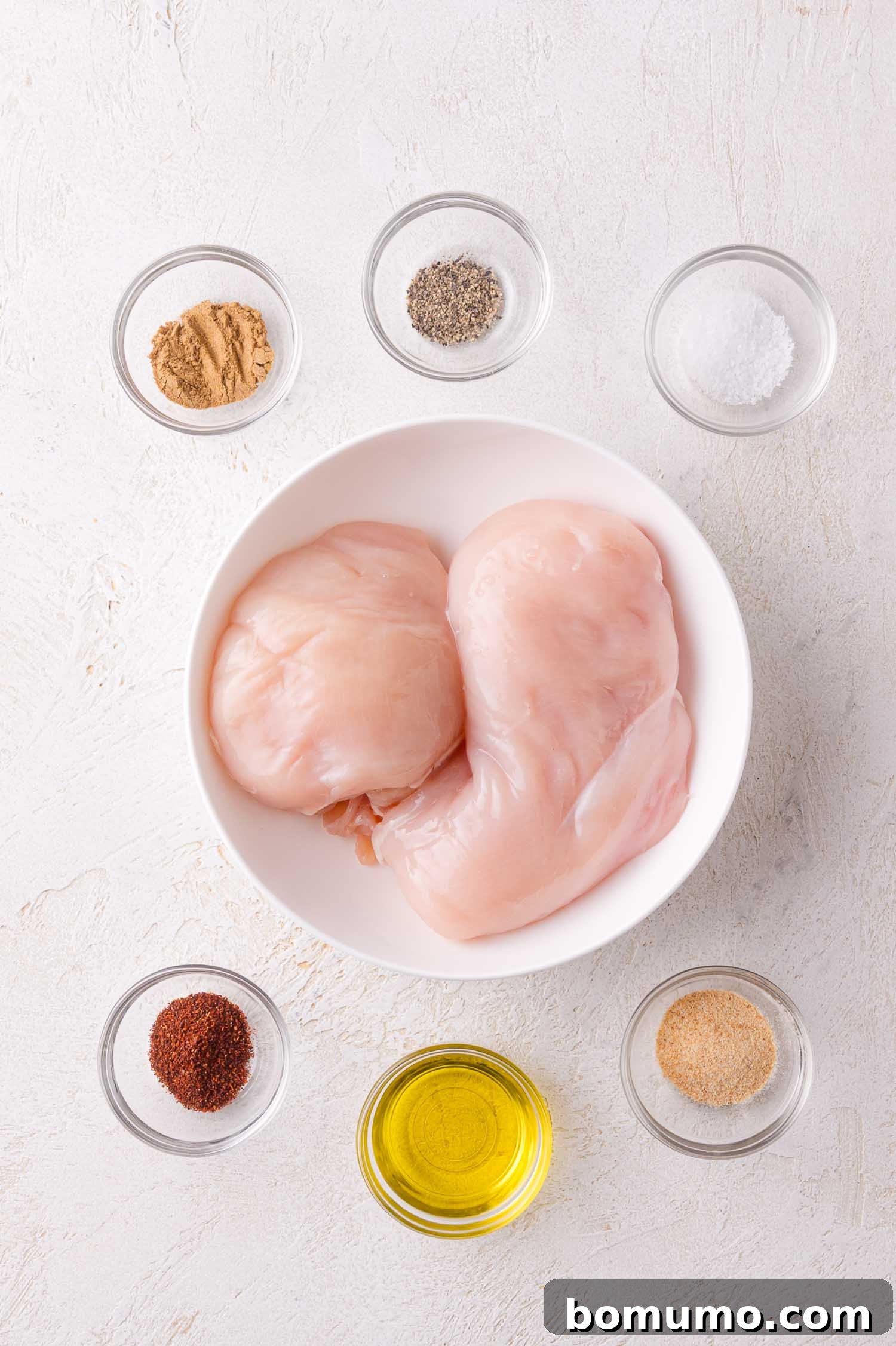 Raw chicken breasts surrounded by small glass bowls containing olive oil, salt, pepper, garlic powder, chili powder, and cumin on a white surface, showcasing the chicken seasoning components.