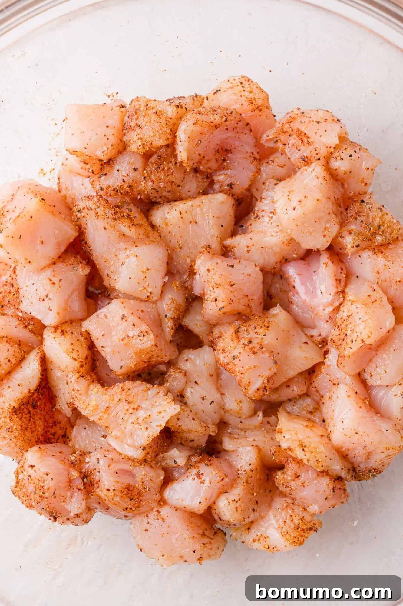 Diced raw chicken pieces, thoroughly coated with a blend of savory seasonings, rest in a clear glass bowl, showcasing their readiness for cooking.
