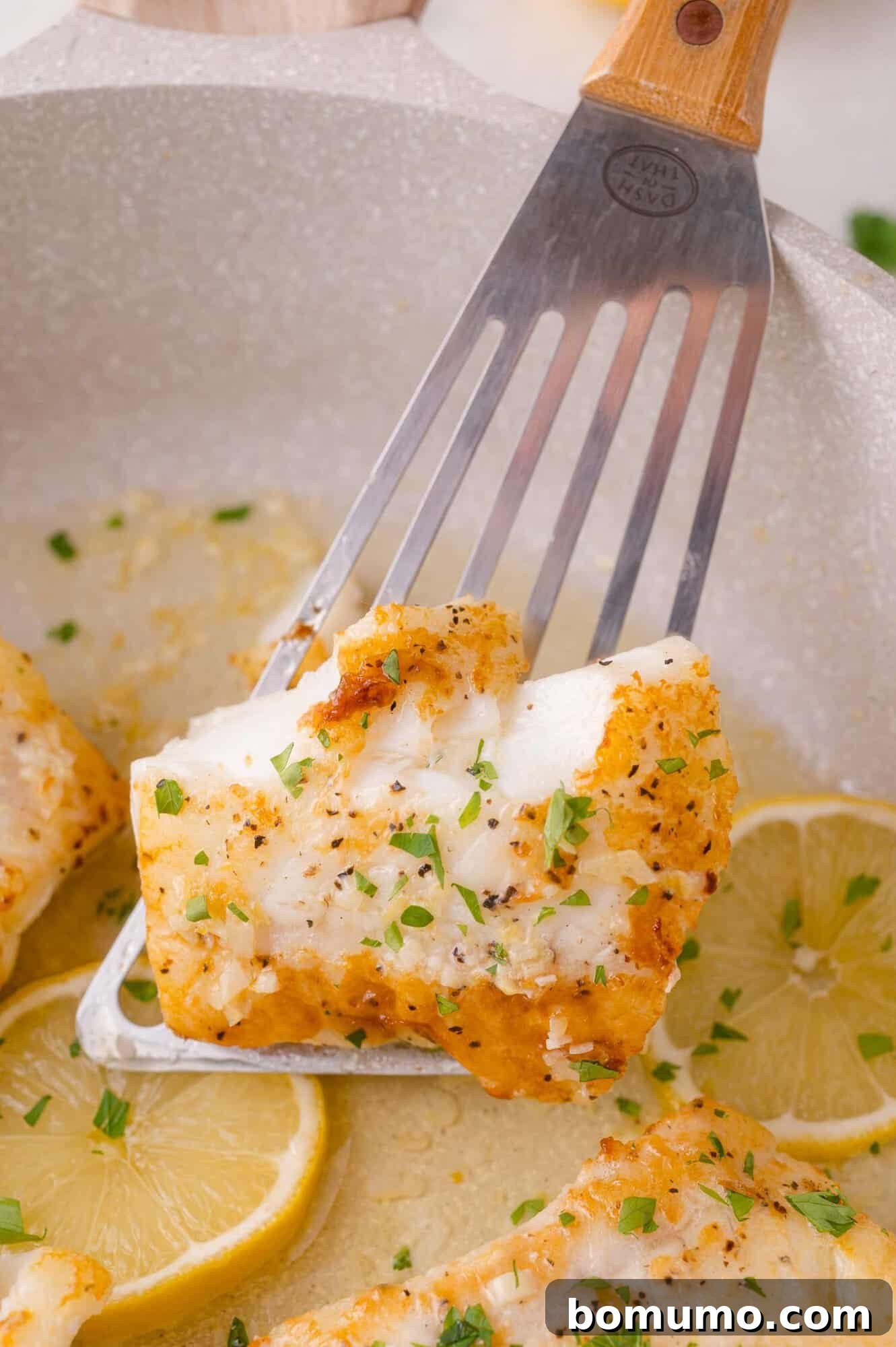Golden Lemon Butter Cod 2 A perfectly seared fillet of lemon butter cod being lifted from a skillet with a metal spatula, with fresh lemon slices and herbs in the background.